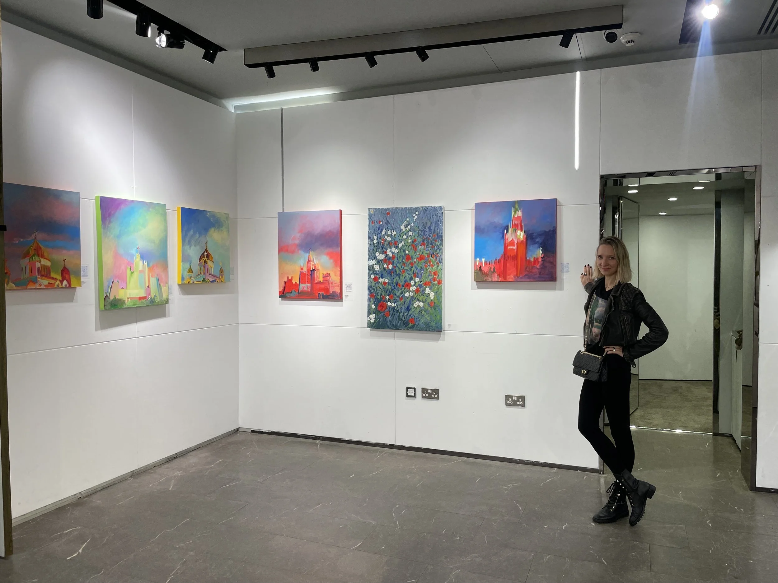 A woman standing in an art gallery next to colorful paintings of buildings and a floral scene on a white wall.