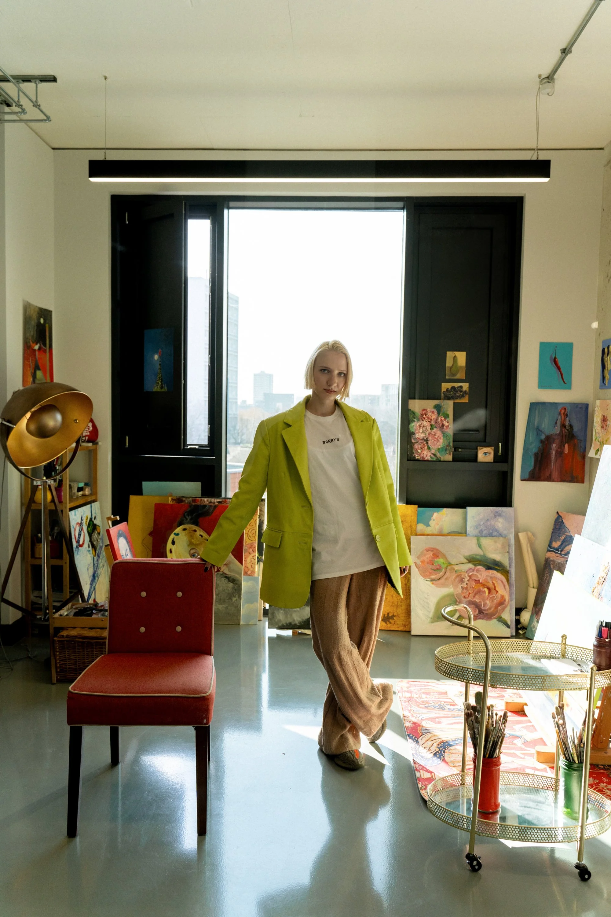 An artist's studio with a woman in a lime green blazer, beige pants, and a white T-shirt standing near a red chair. The studio is filled with colorful paintings, a large window, and art supplies.