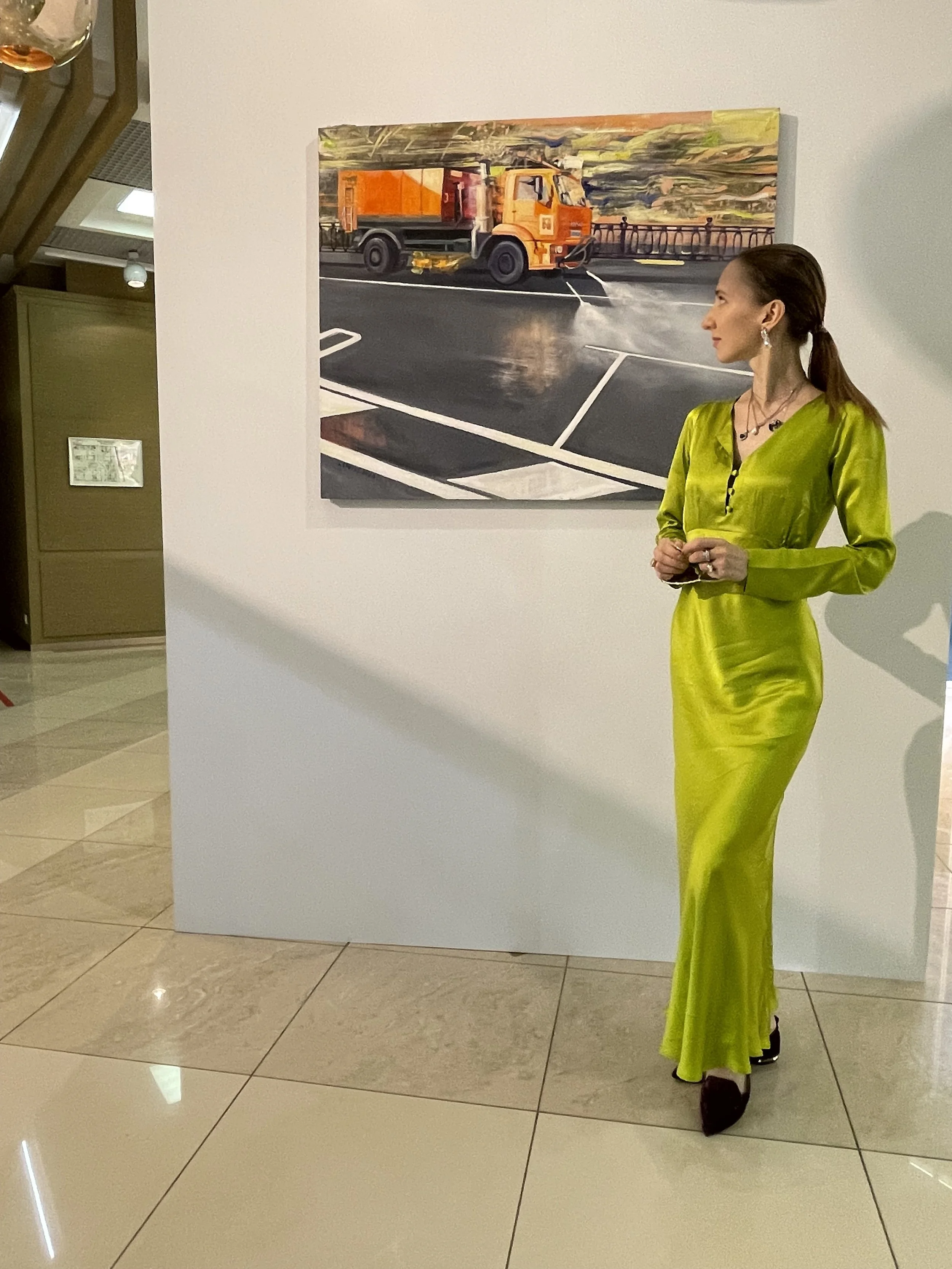 A woman in a bright yellow-green satin dress standing next to a white wall with a colorful painting of a street scene with an orange street sweeper.