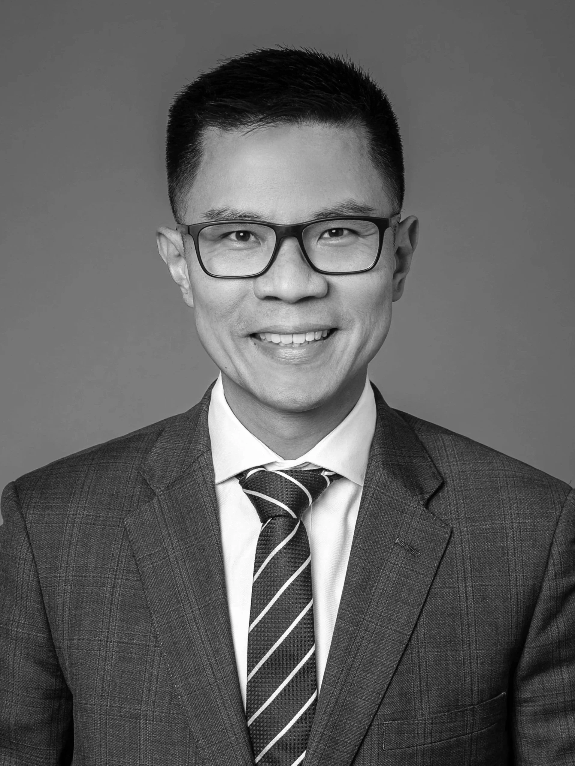 Black and white professional headshot of a smiling man wearing glasses, a suit, shirt, and striped tie against a plain background.
