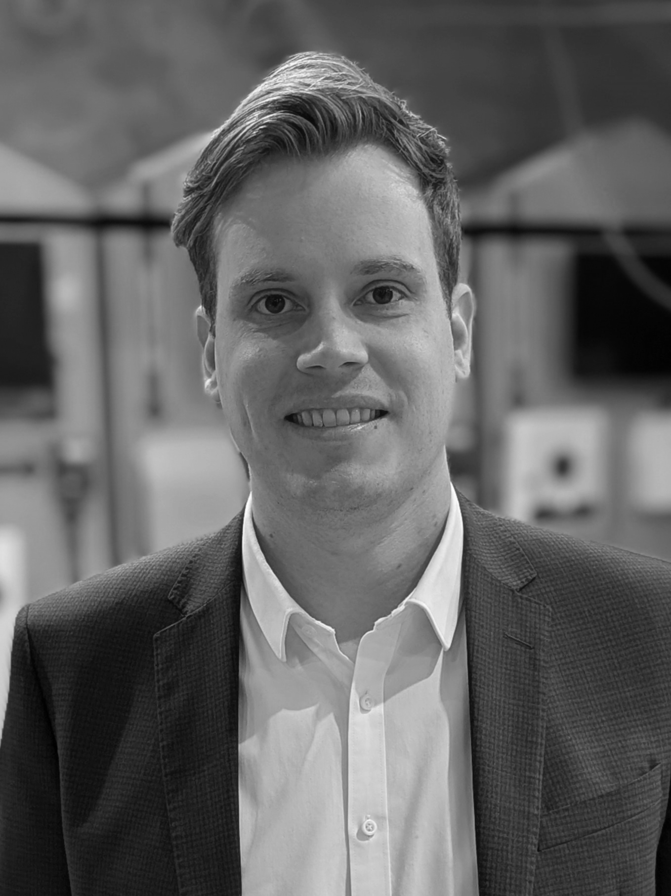 Black and white photo of a young man with short hair, wearing a suit jacket and white shirt, smiling slightly, with an outdoor background.