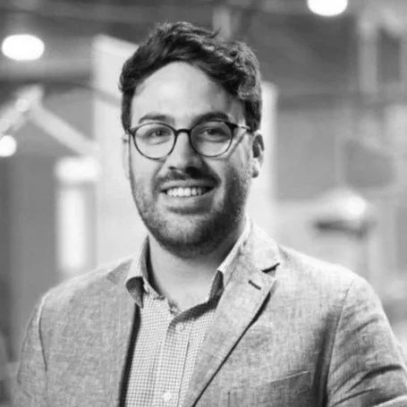 A smiling man with dark hair, glasses, and a beard, wearing a checkered shirt and blazer, standing indoors.