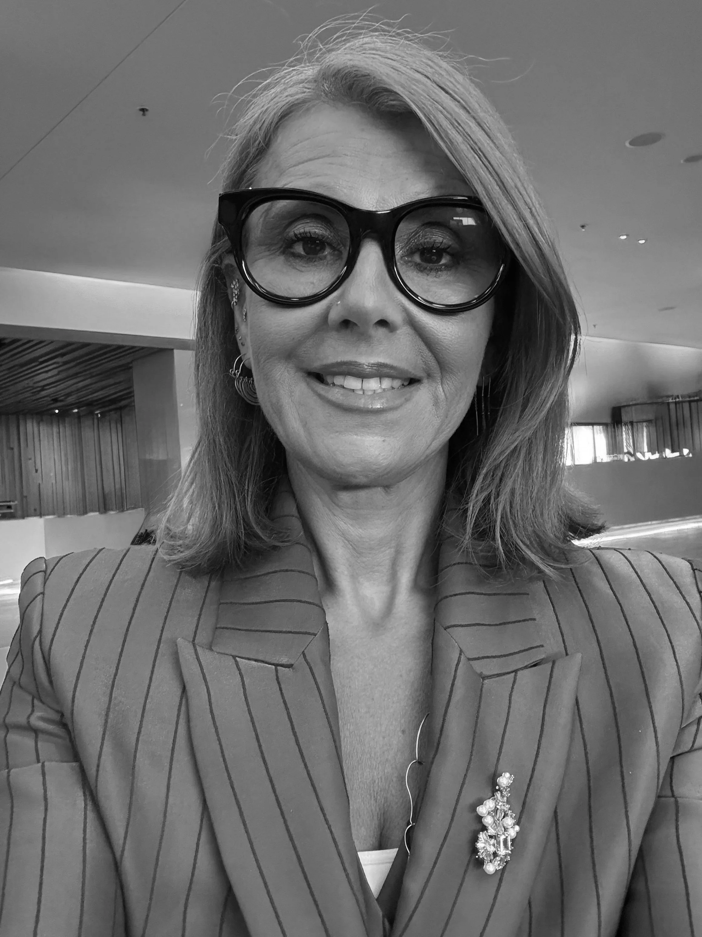A woman with shoulder-length hair, wearing large glasses and a pinstripe blazer, smiles for a selfie in an indoor setting.