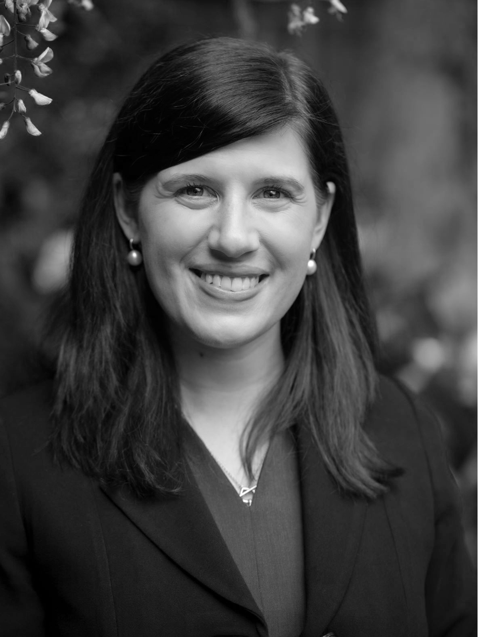 Black and white portrait of a smiling woman with shoulder-length dark hair, wearing earrings and a professional suit, outdoors background.