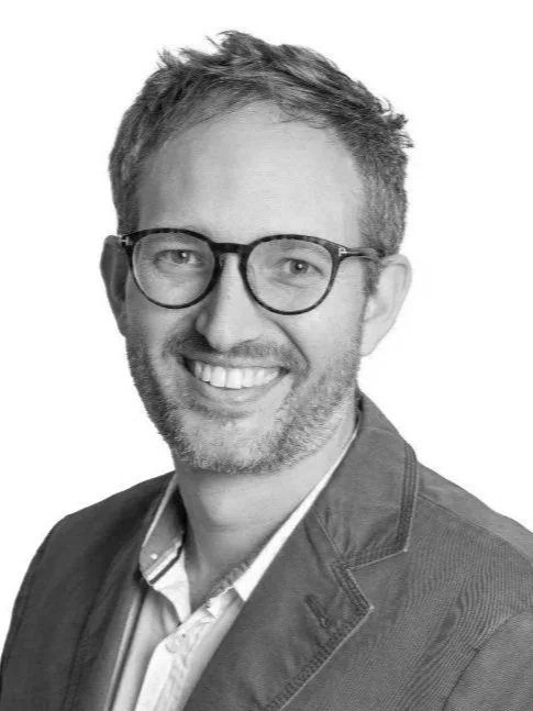 Black and white portrait of a smiling man with glasses, short hair, and a beard, wearing a blazer over a collared shirt.