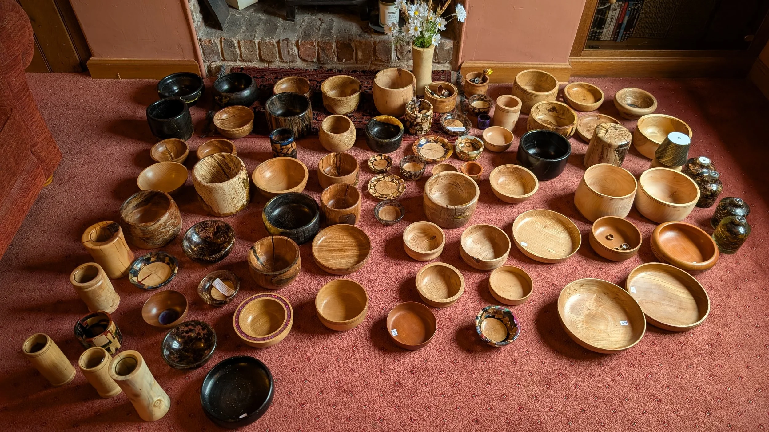 A large collection of wooden bowls of various sizes and colors arranged on a red carpet in front of a brick fireplace and wooden shelving.