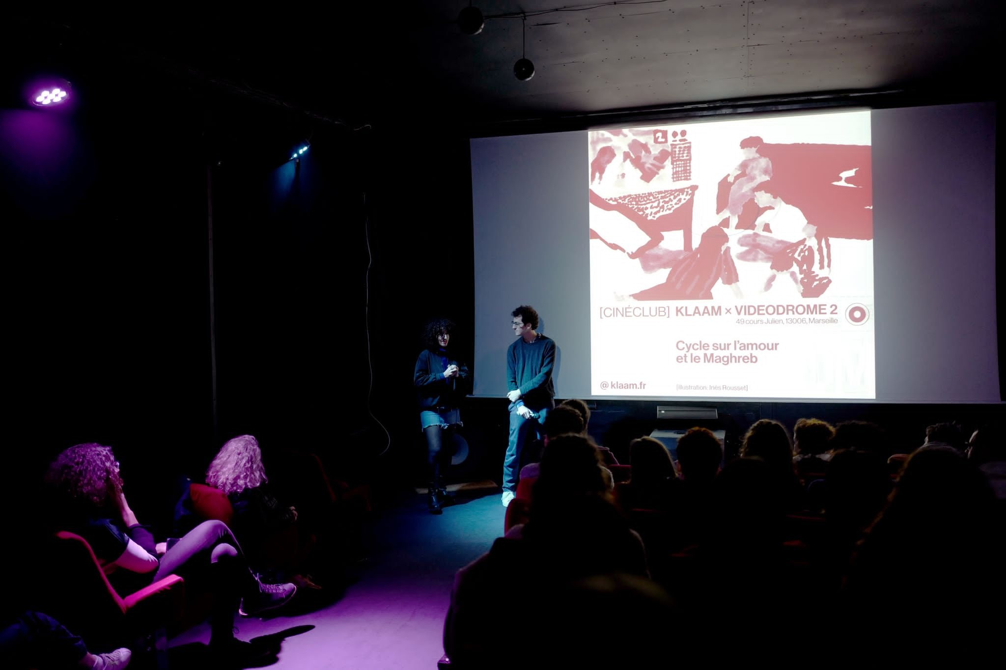 Une présentation ou conférence dans une salle sombre avec deux intervenants devant un écran projetant une affiche promotionnelle en français. Le public regarde la scène, assis dans des fauteuils rouges.