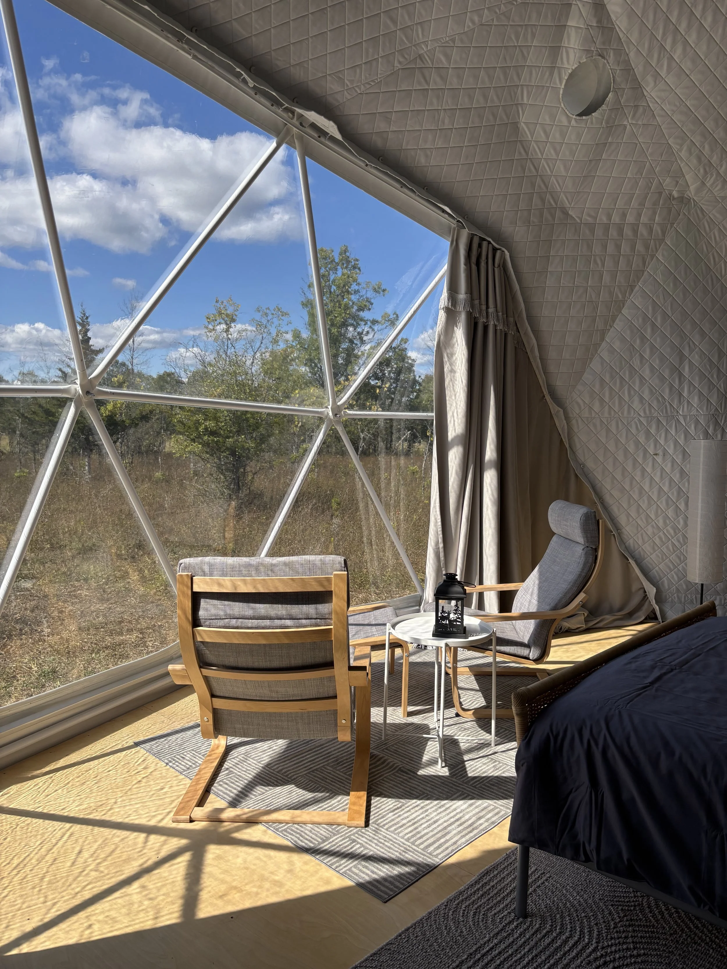A seating area in a white geodesic glamping dome looking out a panoramic window towards a field with cedar trees against the backdrop of a blue sky with white puffy clouds.