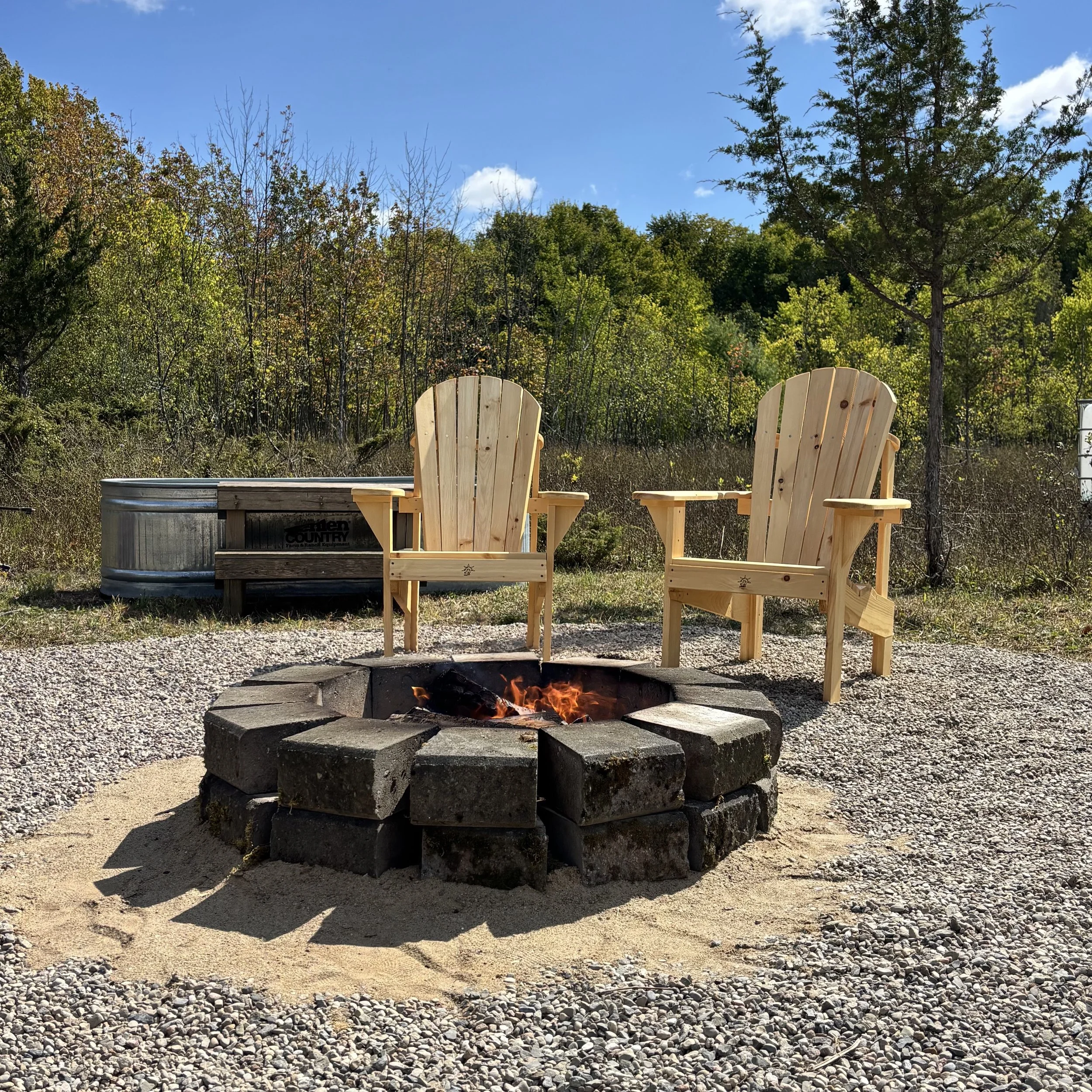 A round stone firepit surrounded by sand and stone. There is a small campfire burning inside the ring of stone. Two pine Muskoka chairs face the campfire and a wood-fired stock-tank hot tub is visible in the background with wooden steps. In the backg