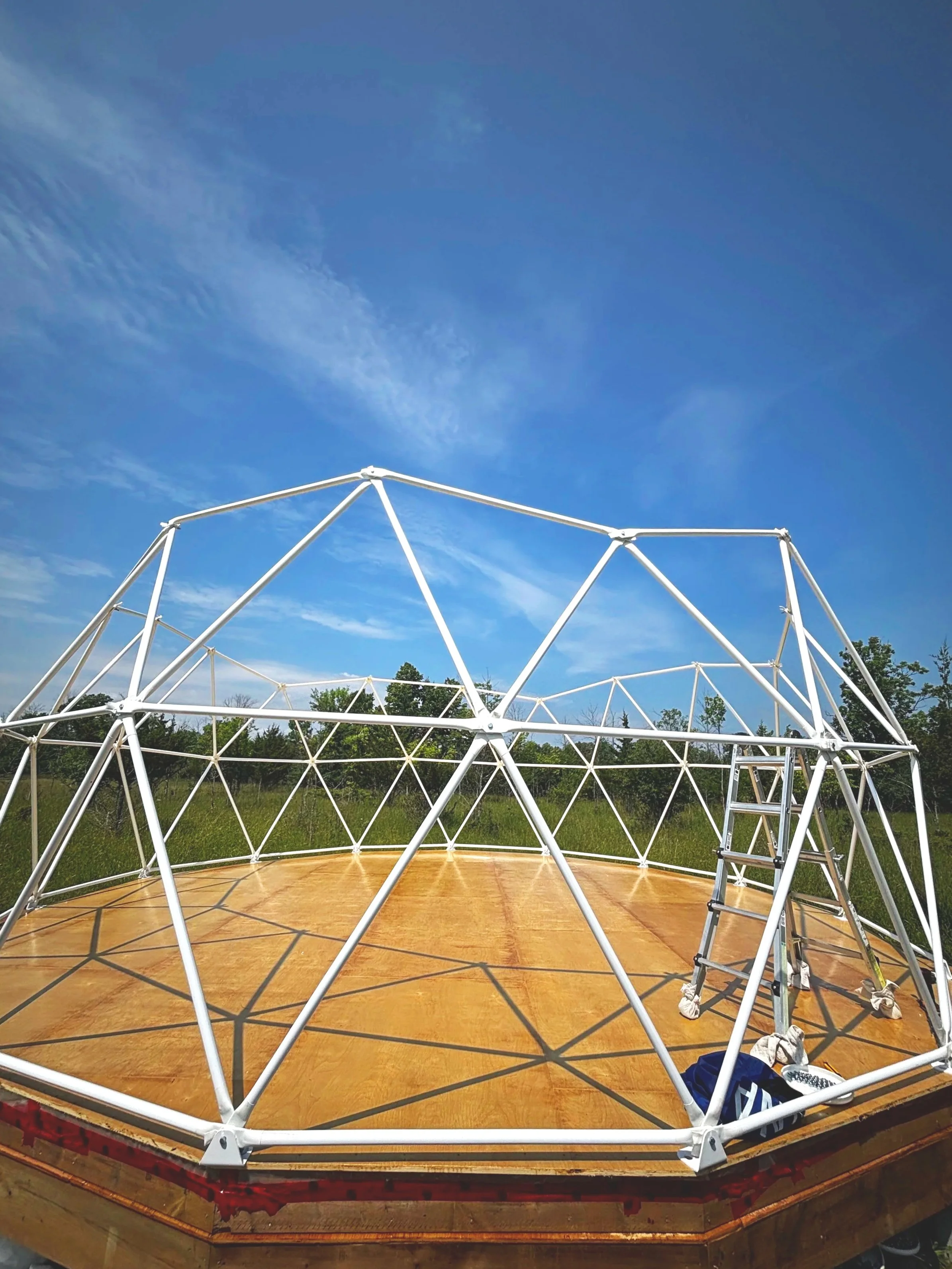 A light brown urethaned wooden polygonal dome platform with white metal bars forming triangular shapes as the beginning of a geodesic dome build. A ladder In the background are green grasses, trees, and a blue sky with white puffy clouds