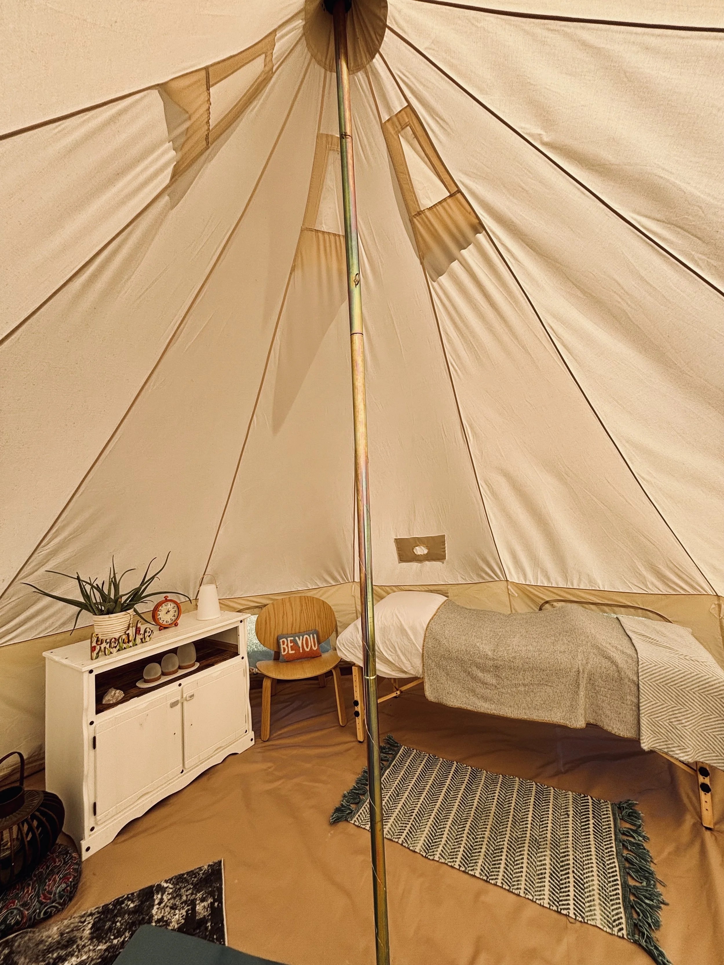The interior of a white canvas bell tent, featuring a massage table draped in linens with a rug beside it. There is also a small chair with a cushion, and a small dresser with a clock, plant and lantern upon it.