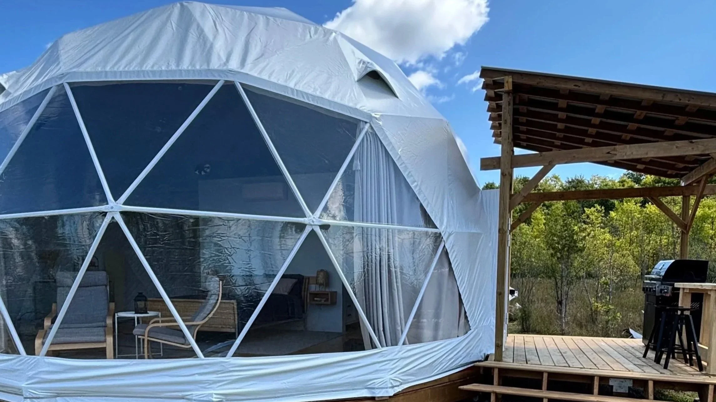 A white geodesic glamping dome, looking in from the outside via a panoramic window. Inside, two IKEA poang chairs are visible, as well as a coffee table. Behind that, inside a queen-sized bed is visible with a floating bedside table with a lamp.