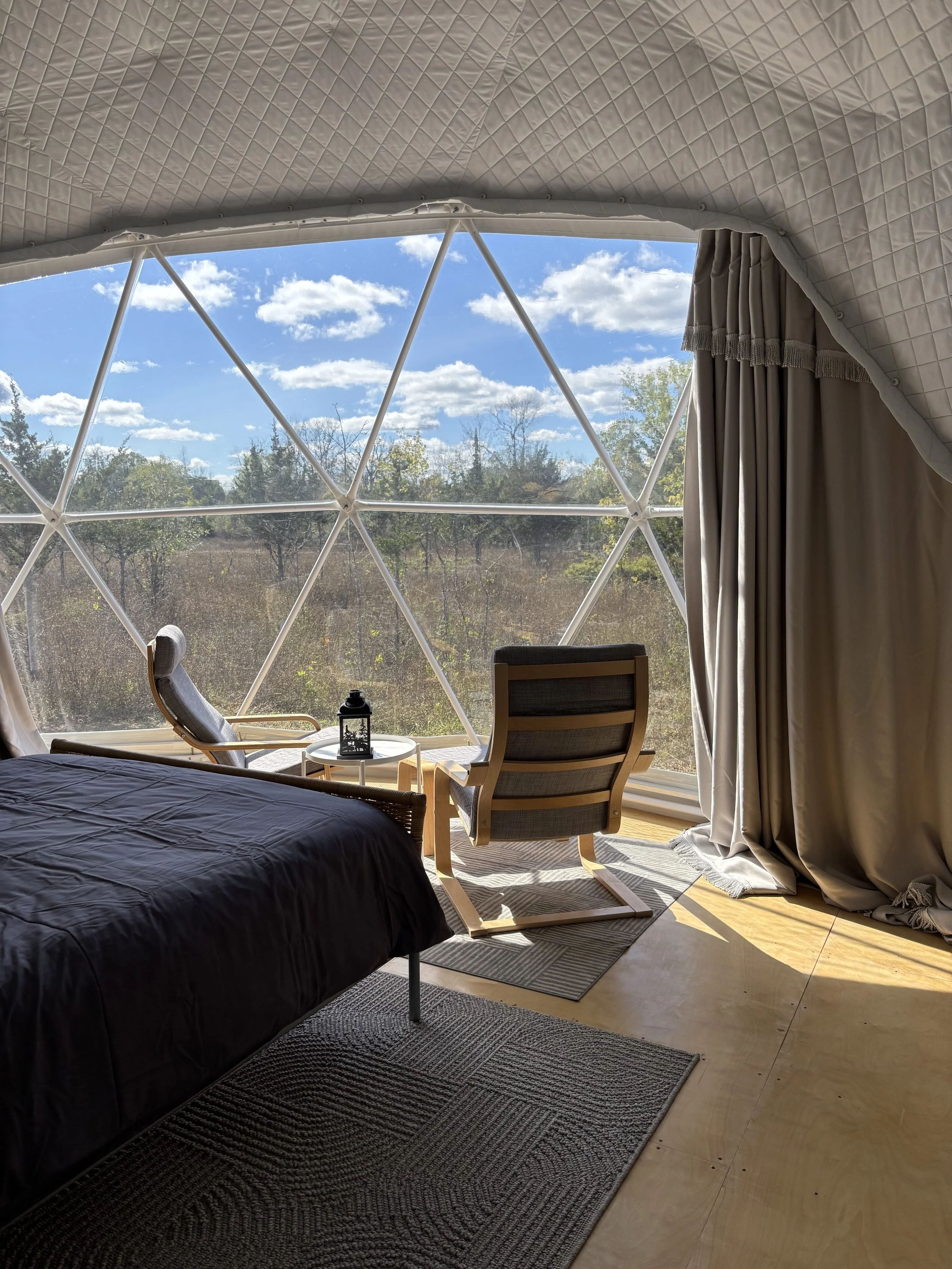 The interior of a white, insulated geodesic glamping dome. The perspective is next to the bed, looking out across the seating area (two IKEA poang chairs and a coffee table on a rug) through the panoramic window. Outside, there are grasses and cedar 