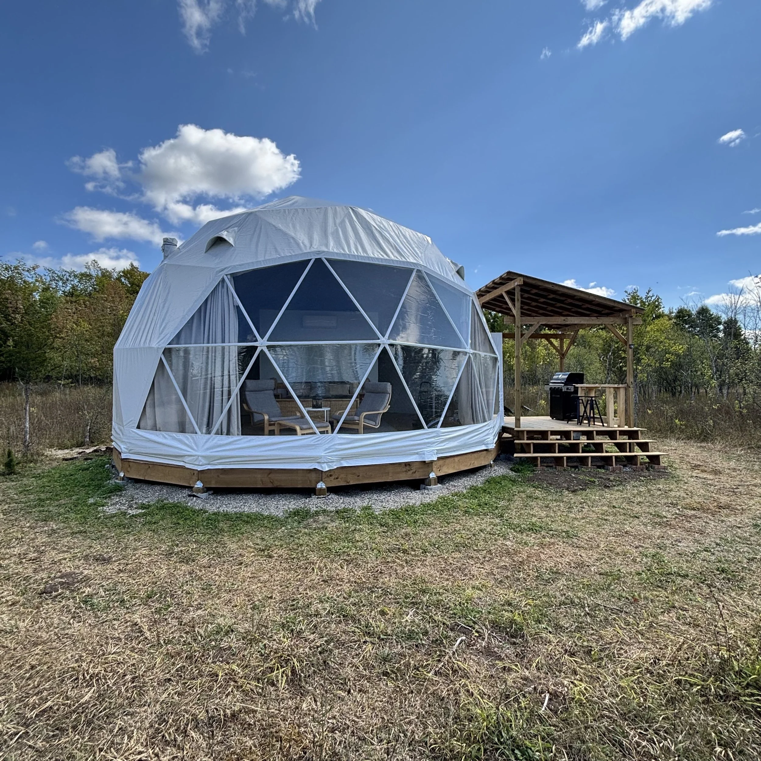 An image of the exterior of a white geodesic dome. A large panoramic window is visible. To the right of the dome is an attached wooden covered deck with a black BBQ, wooden counter and two black stools. The ground is grassy. In the background are gra