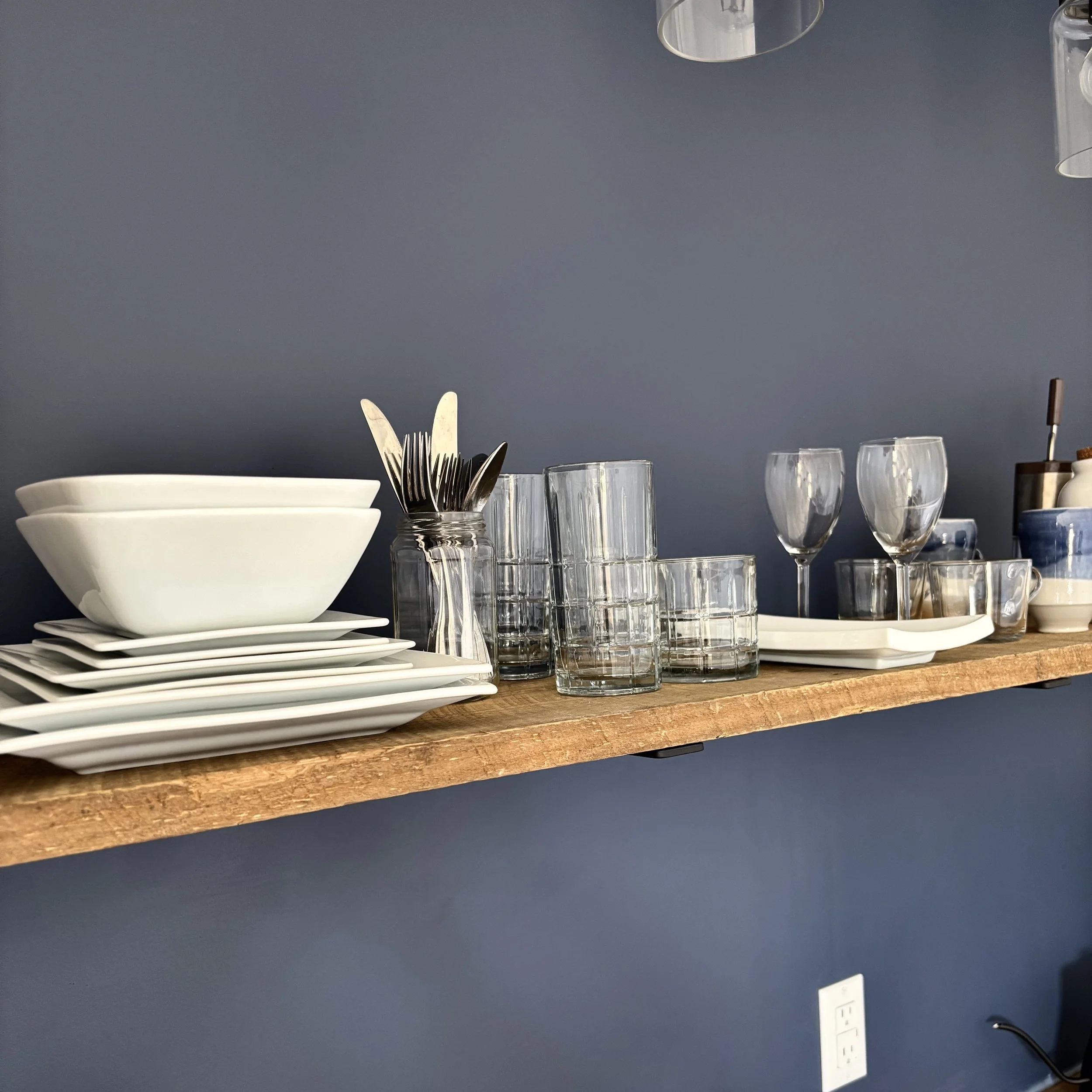 A close-up of white dishes and glasses as well as cutlery on a wooden floating shelf against a navy blue wall. 