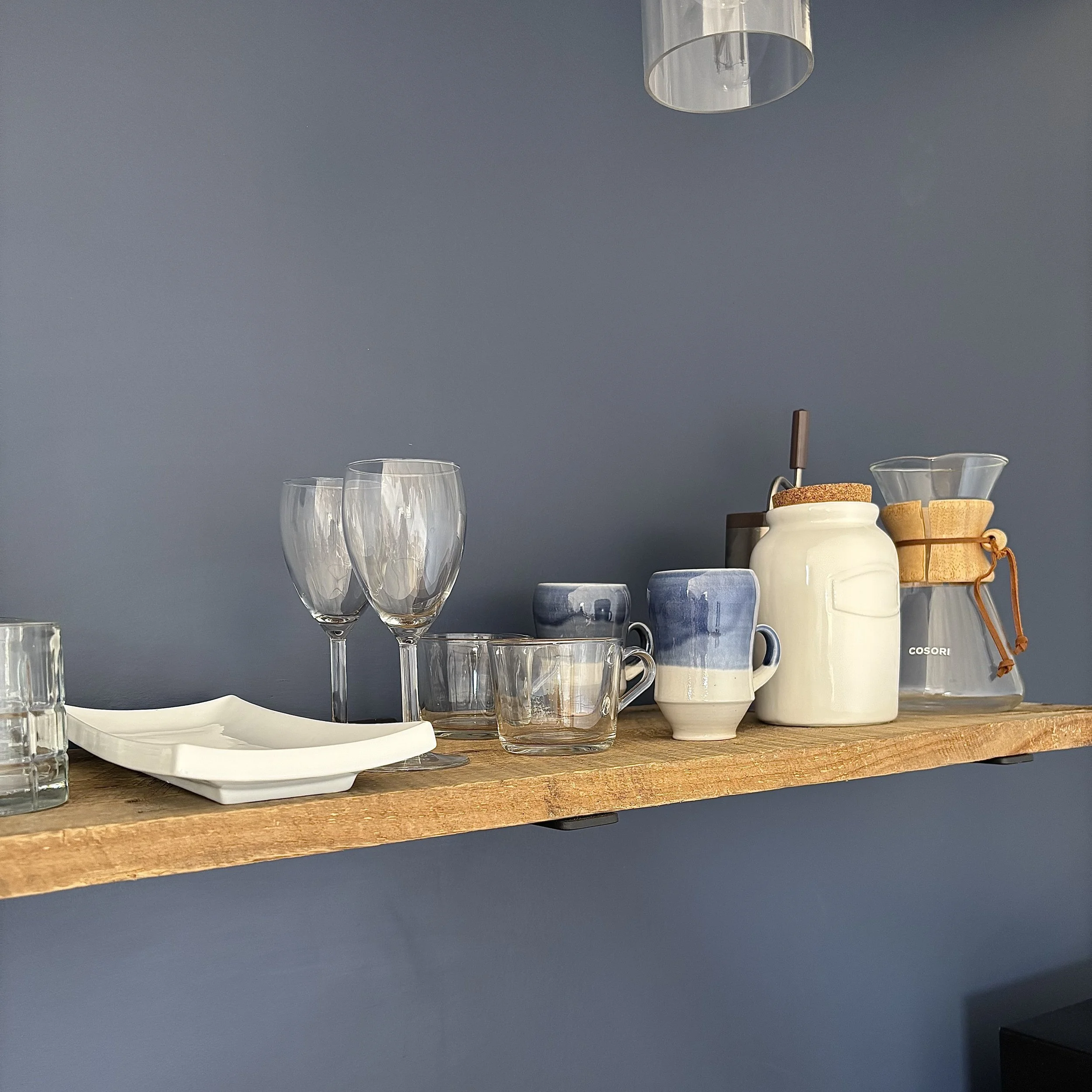 A close-up of white dishes and glasses as well as a pour-over coffee set on a wooden floating shelf against a navy blue wall. 