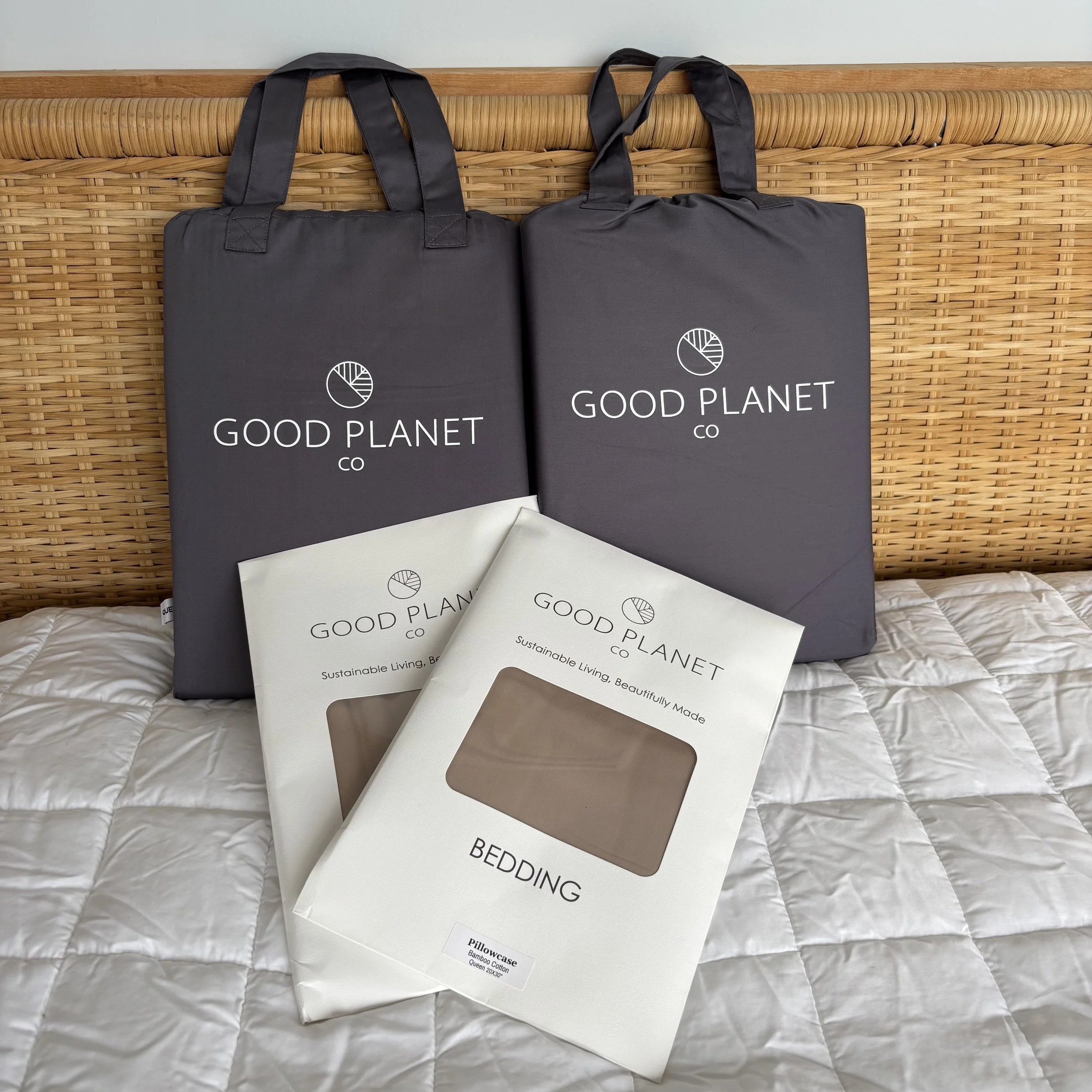 An unopened charcoal grey sheet set, duvet cover and two paper envelopes of dune-coloured pillowcases sitting on a mattress cover against a rattan headboard.
