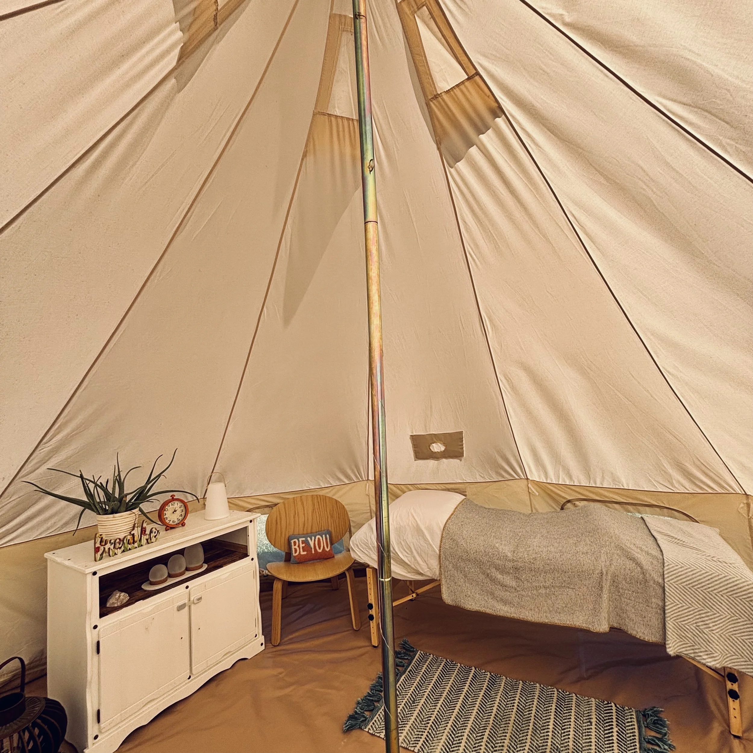 The interior of a cream-coloured canvas bell tent. Inside is a white cabinet with a plant, analog clock and some lamps. There is a wooden accent chair with a throw cushion that says "Be You." There is a massage table with white sheets & grey blankets