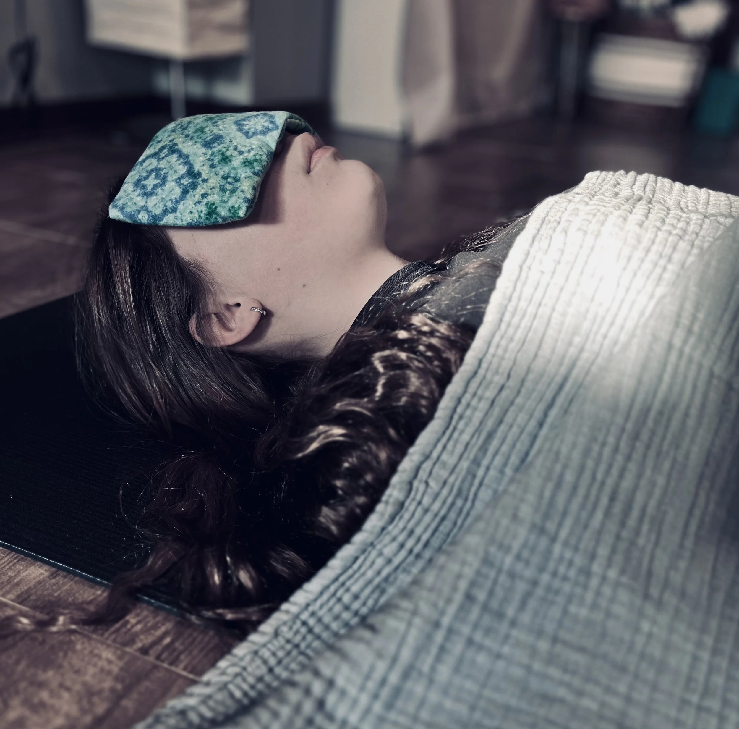 A white female with brown hair lying on a yoga mat. She is draped with a blue blanket on her body and a blue eye pillow over her eyes.