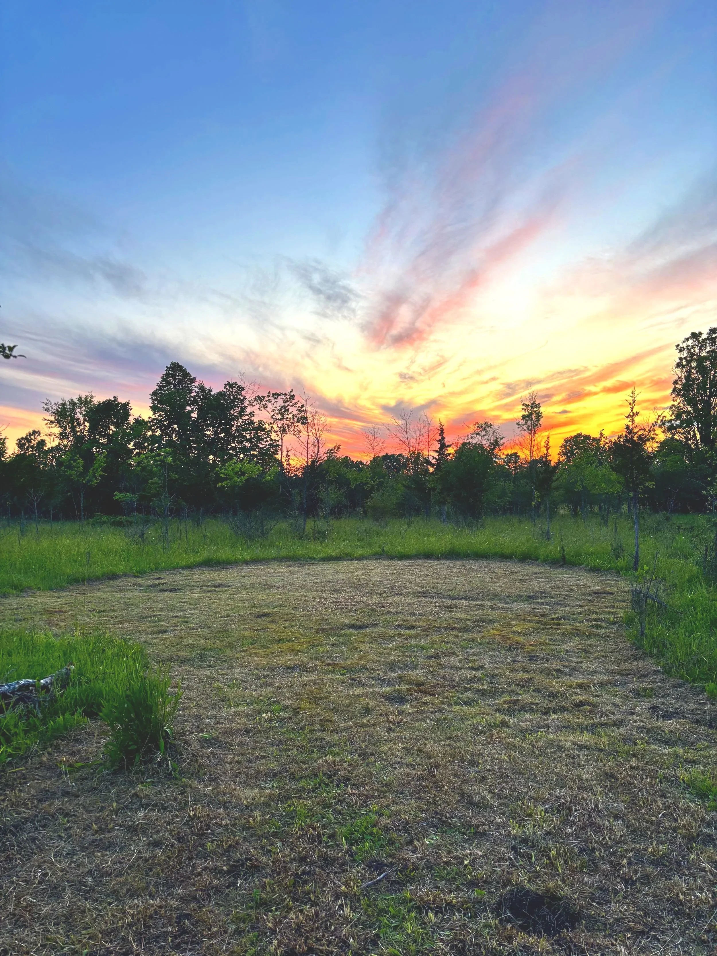 A green, grassy area, with a large circle where the grass has been cut short. Around the circle are green grasses and trees. The sun is setting and the sky is a mixture of blues, purples, oranges and pinks streaked with clouds.