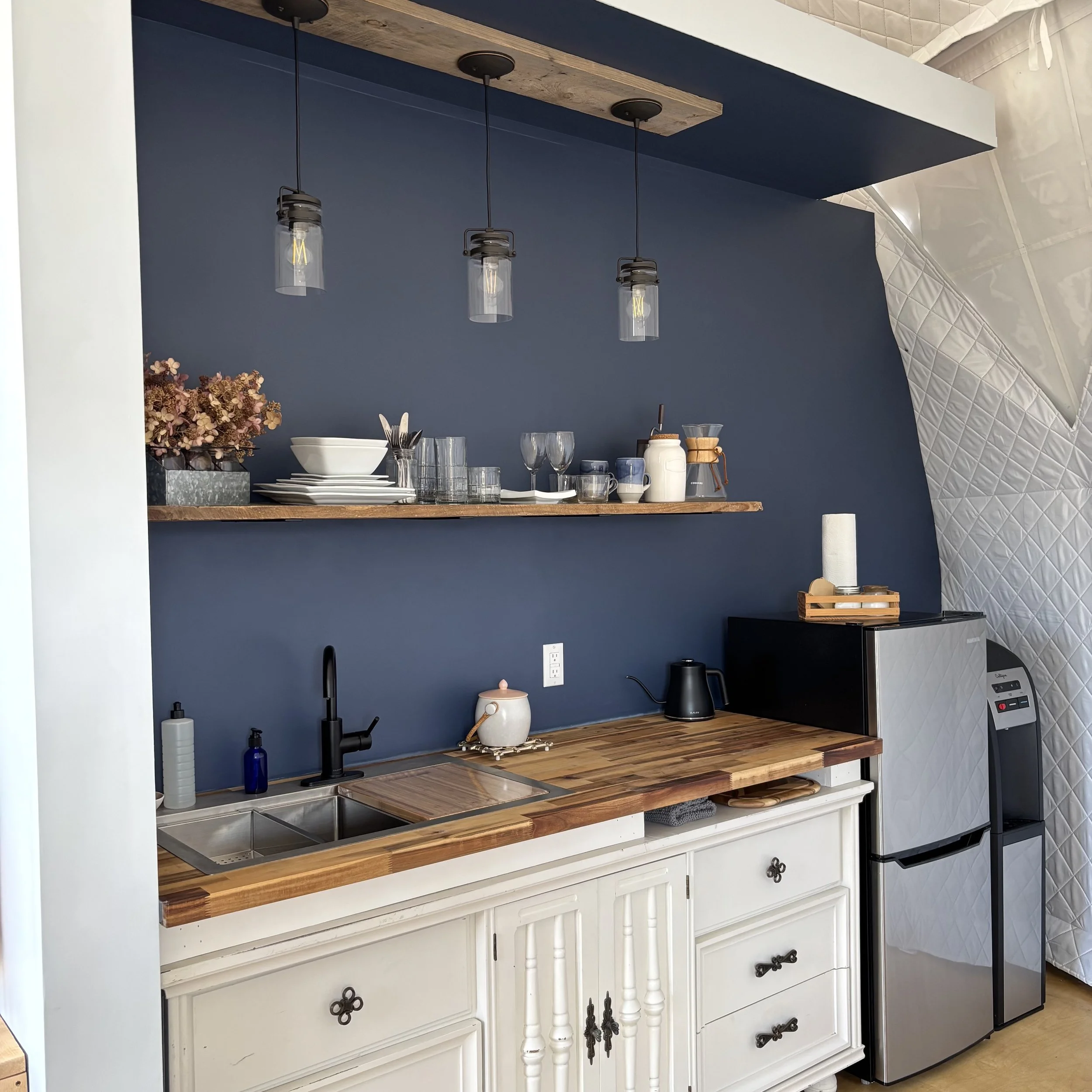 A kitchenette in a white insulated geodesic glamping dome. There are three glass pendant lights showcasing a wood floating shelf holding dried flowers, glasses and white dishes as well as a pour-over coffee set. The countertop is butcher block over a