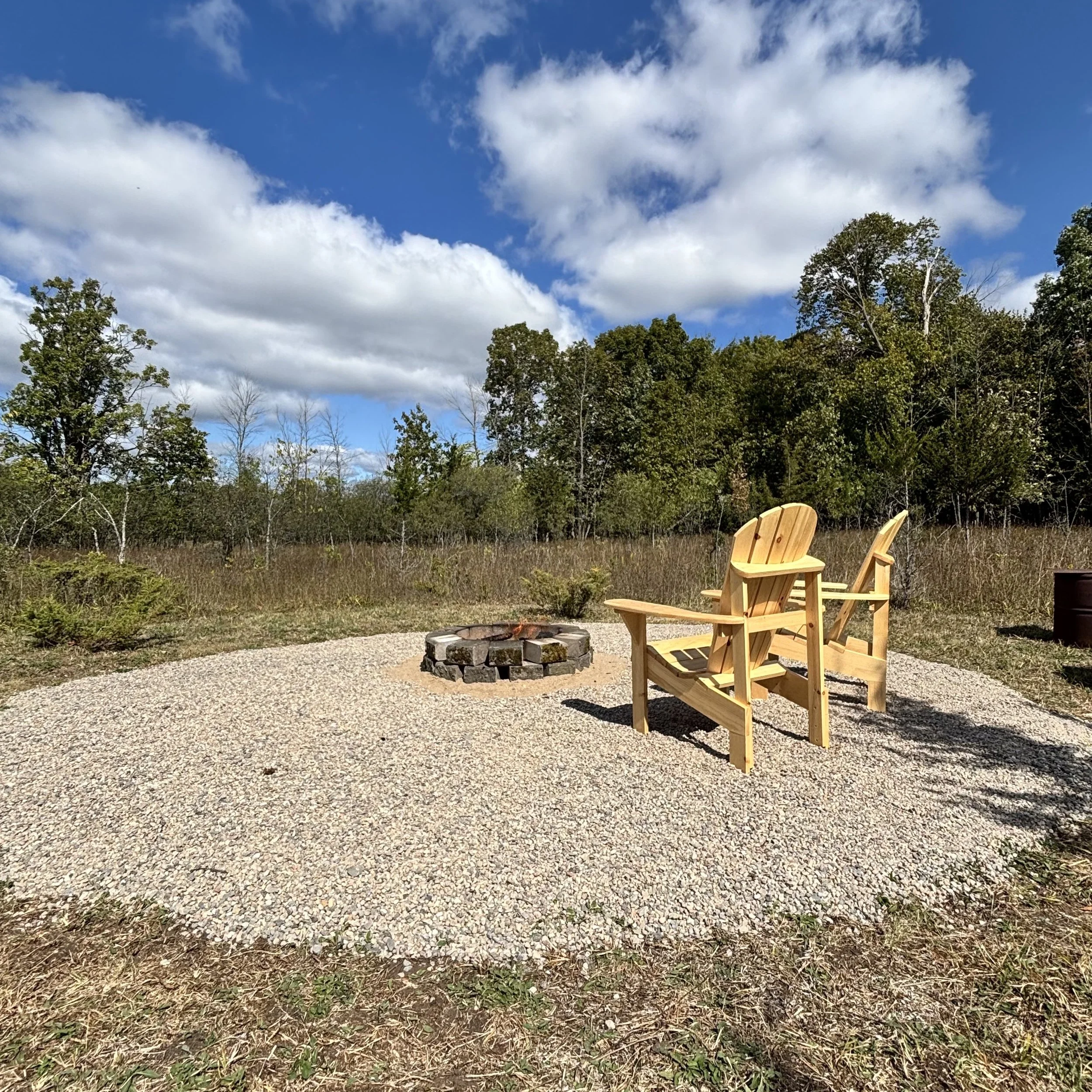 A round stone firepit surrounded by sand and stone. There is a small campfire burning inside the ring of stone. Two pine Muskoka chairs face the campfire. In the background is a juniper bush and grasses. Further back are deciduous trees and a blue sk