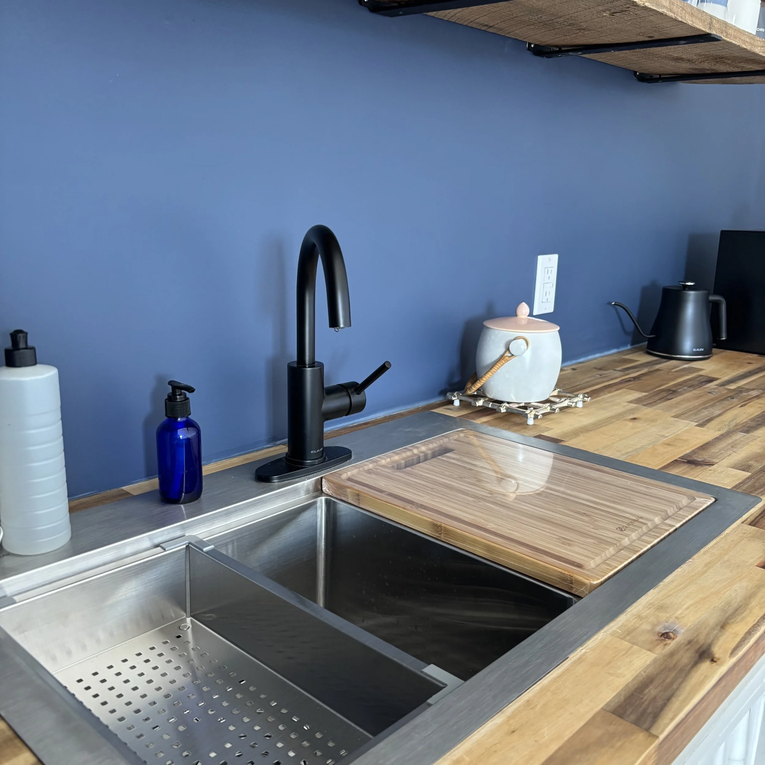 A close-up of a stainless steel double kitchen sink with removable custom stainless steel strainer and wooden cutting board. The faucet is black. There is a bottle of dish soap and a blue glass pump bottle of liquid hand soap. The counter is wooden. 