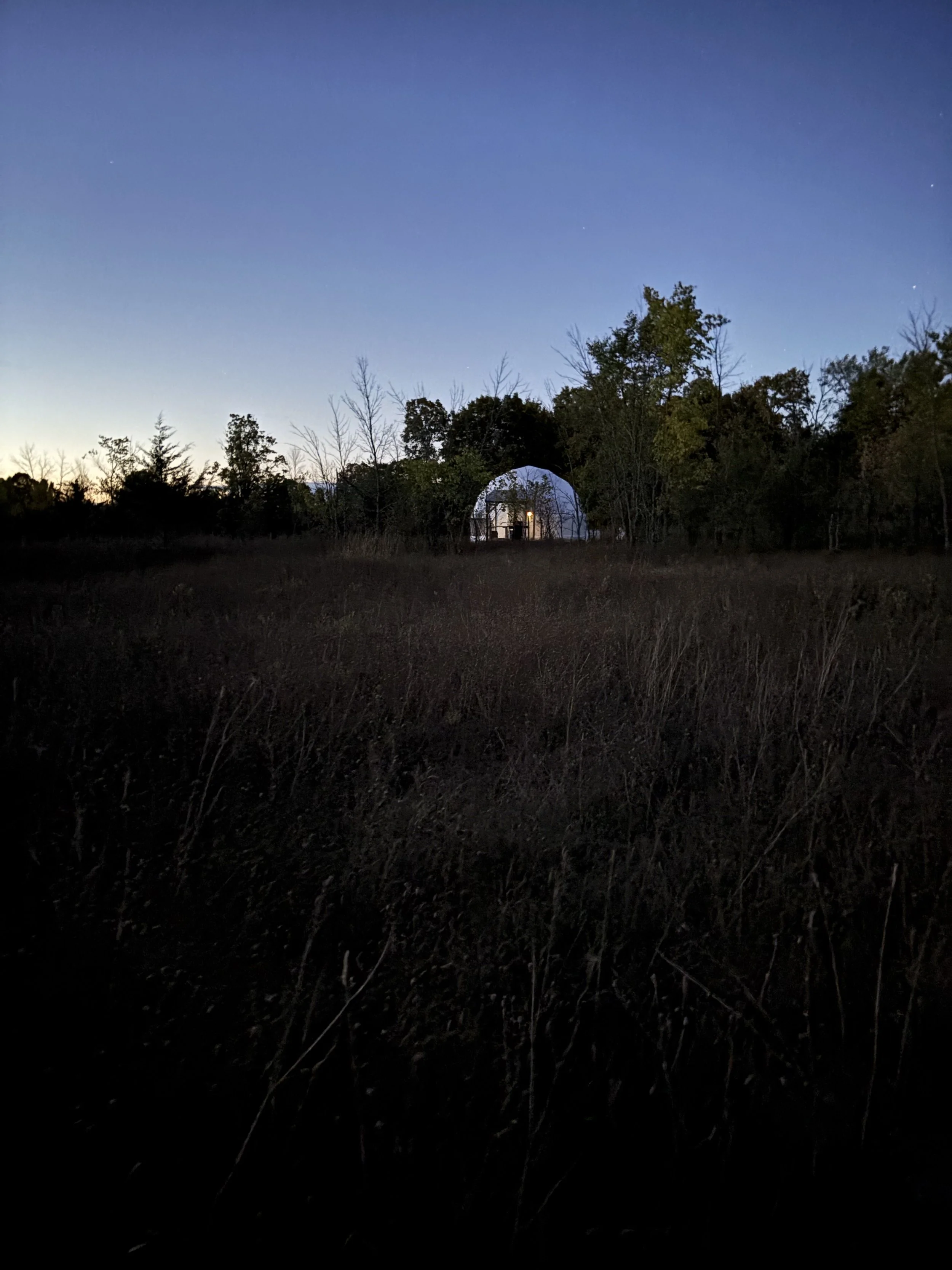 An evening shot from a distance of a white geodesic glamping dome with interior lights on amid silhouetted trees and a blue sky at sundown.