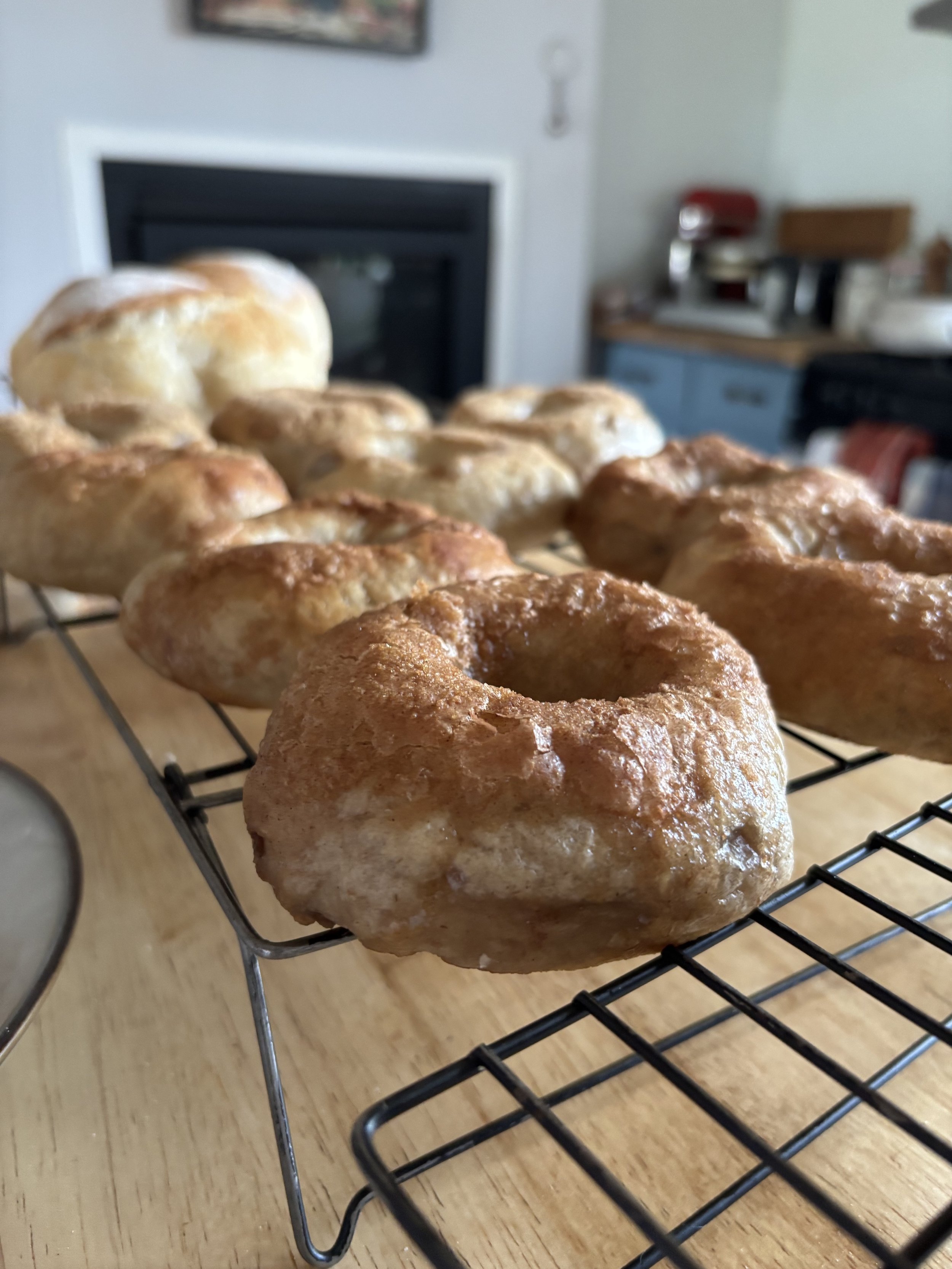 A bunch of apple cinnamon bagels on a baking rack (with sourdough boules in the background) over a wooden countertop. 