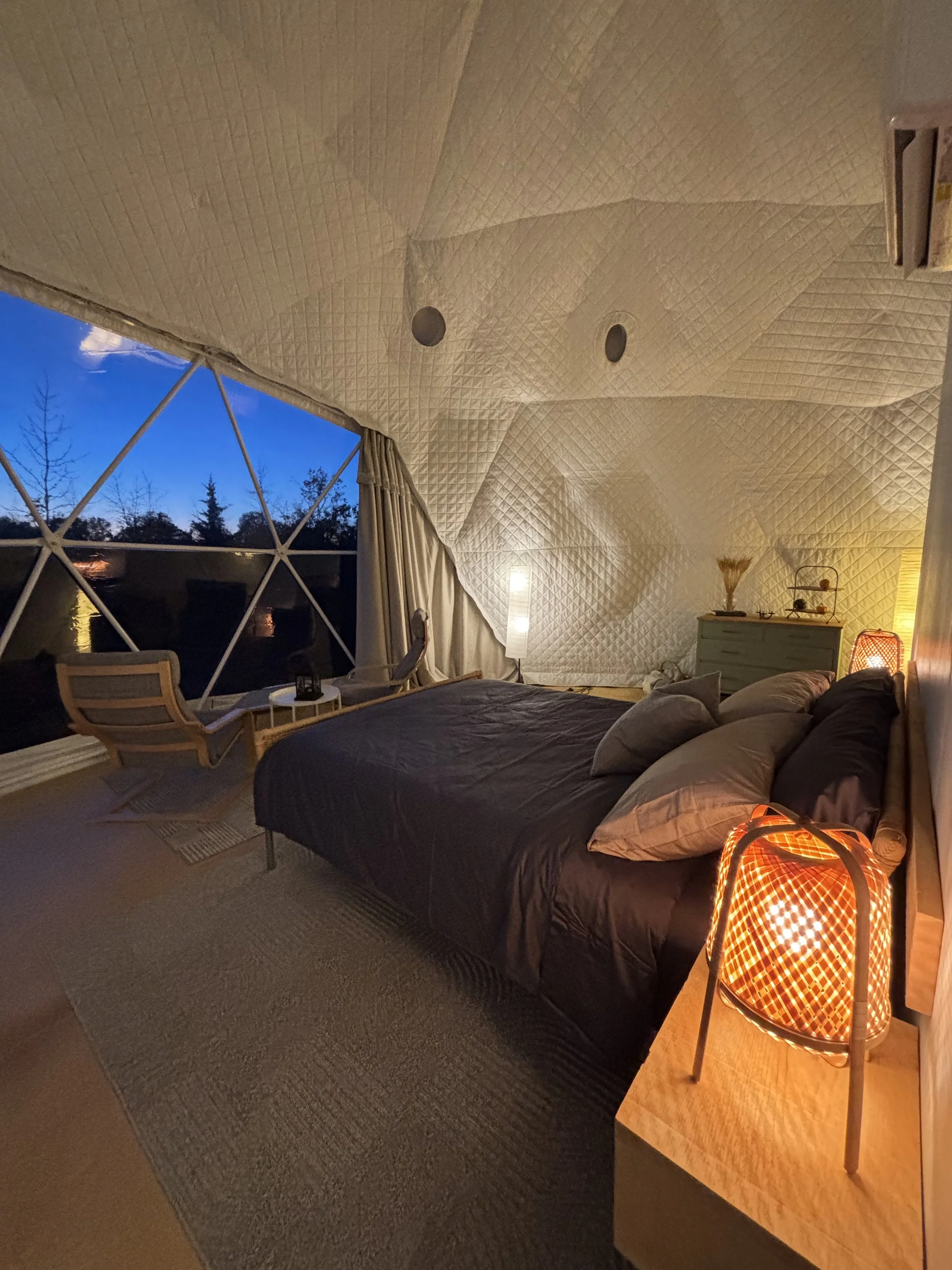 An evening shot of the interior of a white insulated geodesic glamping dome. In the foreground is the warm glow of a basket-woven bedside lamp. A queen-sized bed with charcoal grey sheets is visible, as well as a seating area in front of a panoramic 