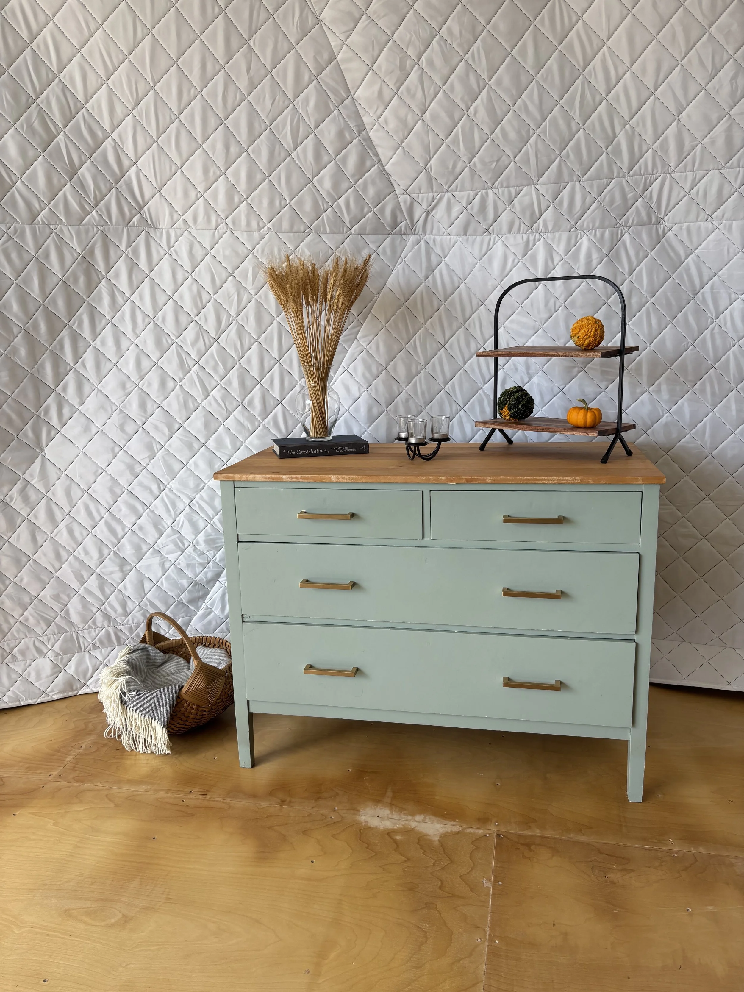 A seafoam green 4-drawer dresser with a wooden top on a light plywood floor in a white insulated geodesic glamping dome. Beside the dresser is a large wooden basket with grey throw blankets. Atop the dresser is a dark blue hard cover book with a glas