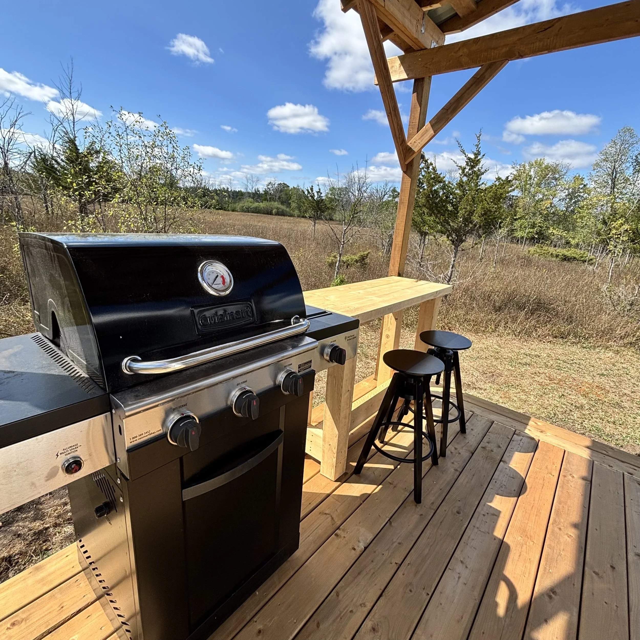 A black BBQ on a wooden covered deck beside a wooden counter with 2 black stools. In the background are grasses and trees and a blue sky with white puffy clouds.