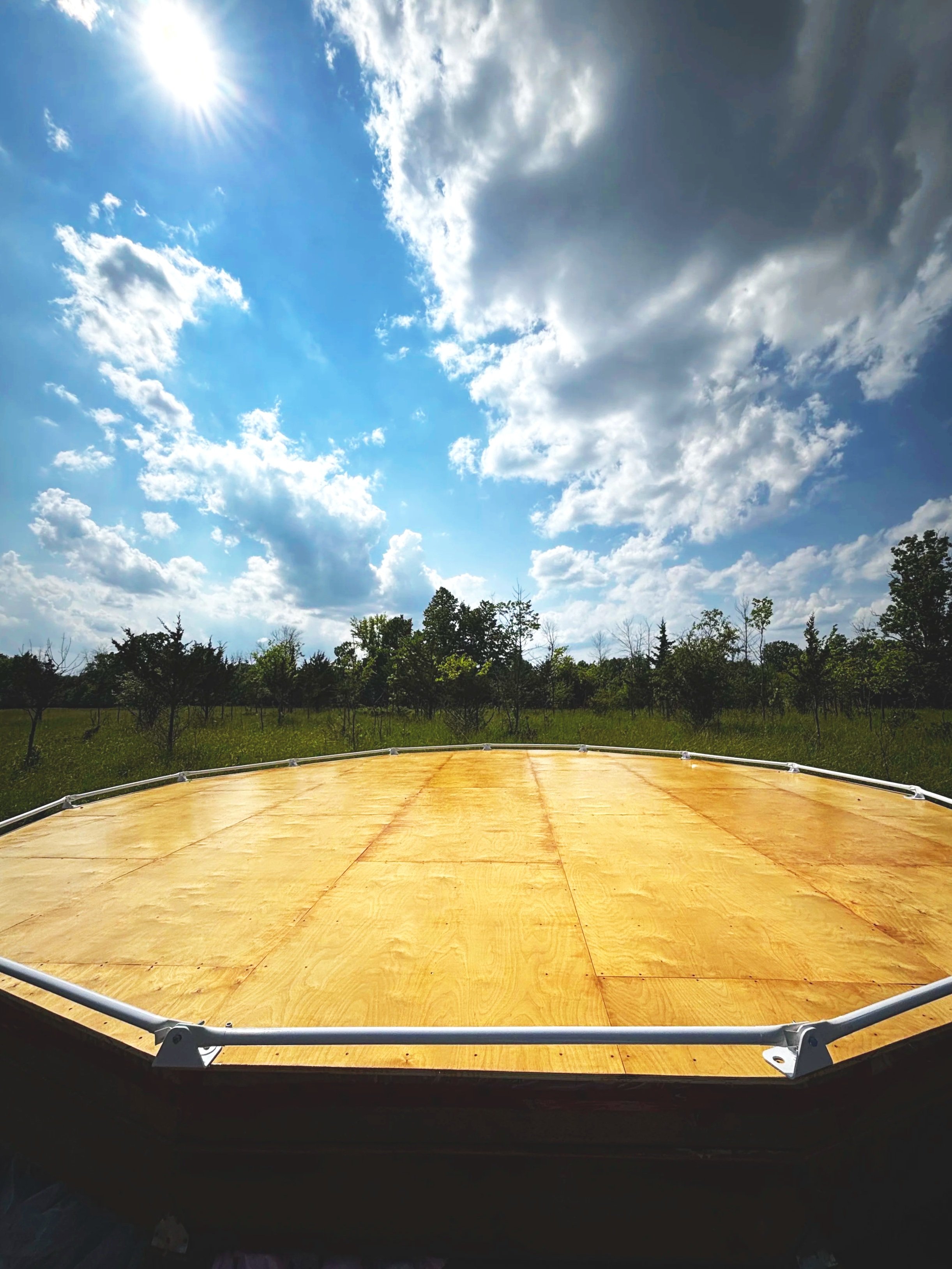 A light brown urethaned wooden polygonal dome platform with bottom white rails installed. In the background are green grasses, trees, and a blue sky with white puffy clouds.