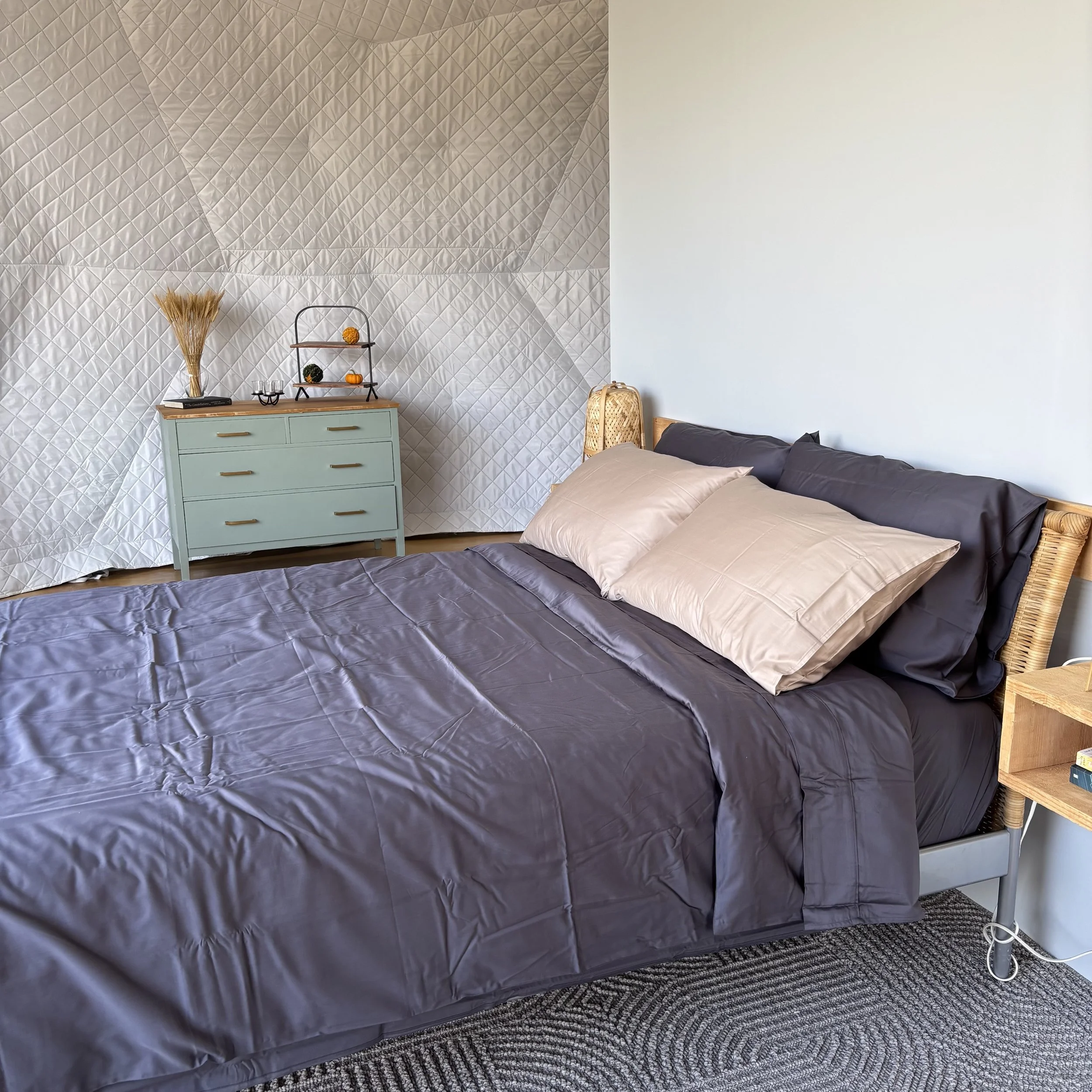A queen-sized bed with rattan headboard and charcoal grey sheets and dune-coloured pillow cases in a white insulated geodesic glamping dome. A grey rug is beneath the bed.