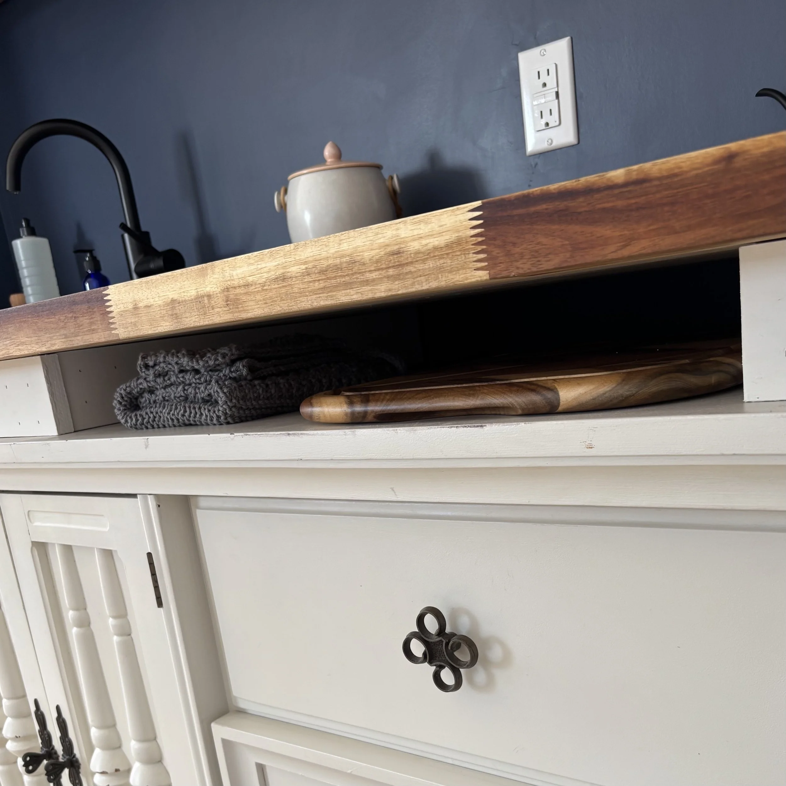A close-up of white cabinetry beneath a wooden counter. There is a wide, thin cubby between the cabinetry and the countertop where a wooden acacia cutting board is stowed as well as a set of knitted grey tea towels and dish cloths. On the counter a c