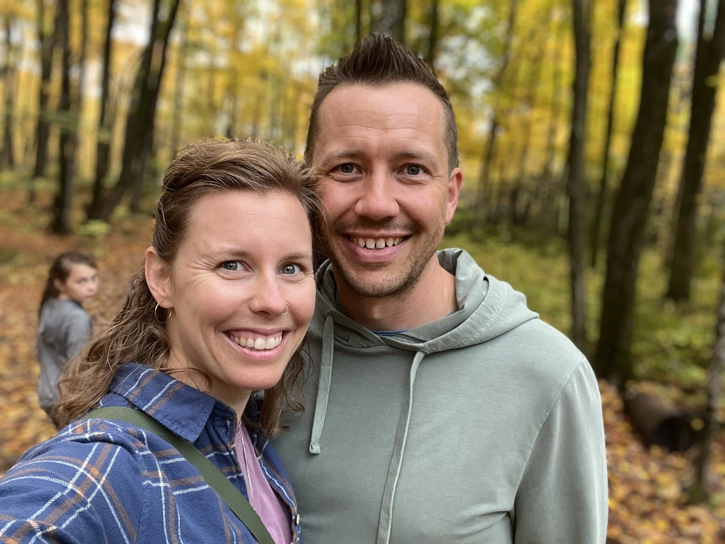 A woman and man with light skin and blue eyes smiling on a trail in a forest.