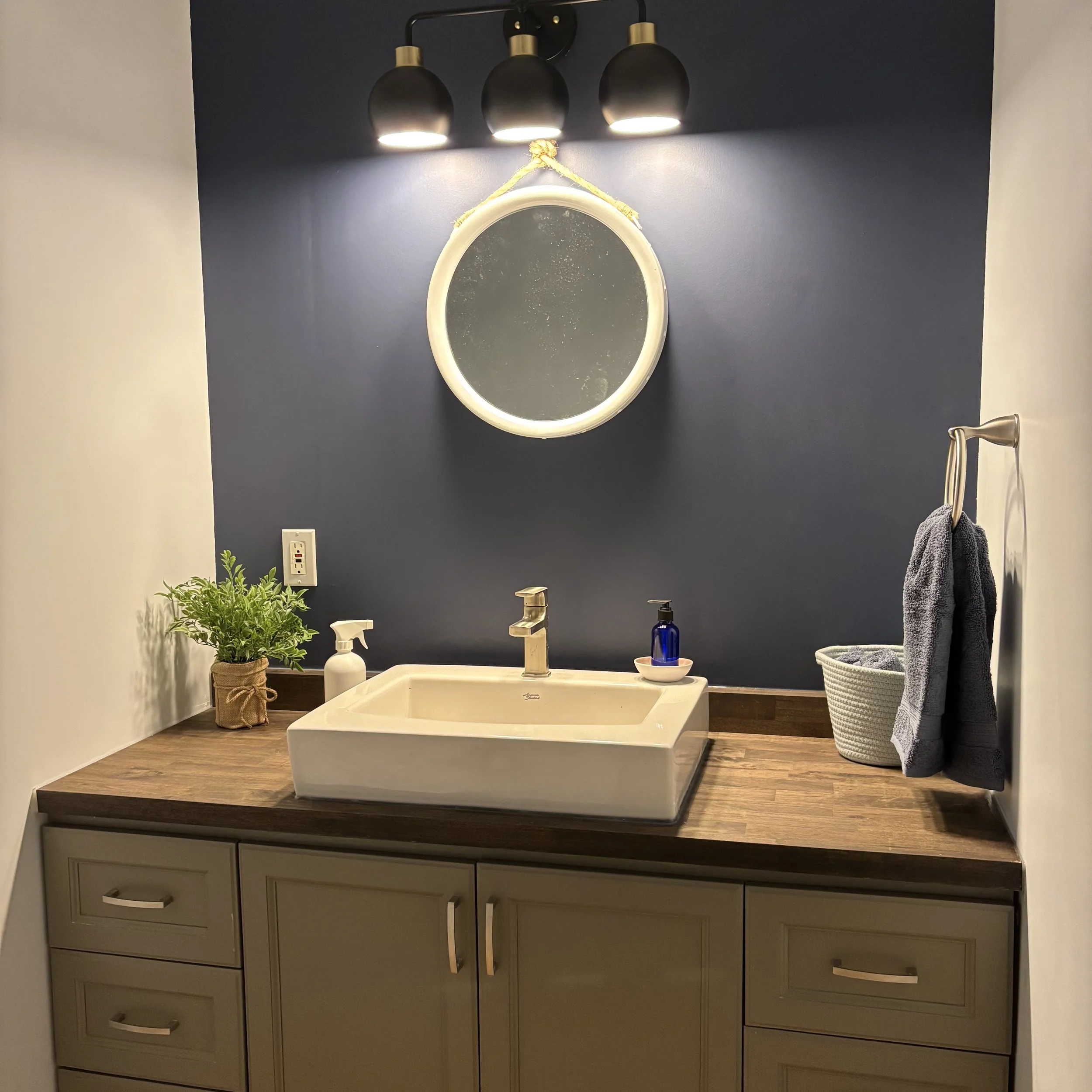 A bathroom vanity with dark grey/green cabinetry and a dark wood countertop. Atop the counter sits a white bathroom sink. Beside the sink is a plant, a white spray bottle, as well as a blue rope basket. A blue glass hand soap pump is visible on the s