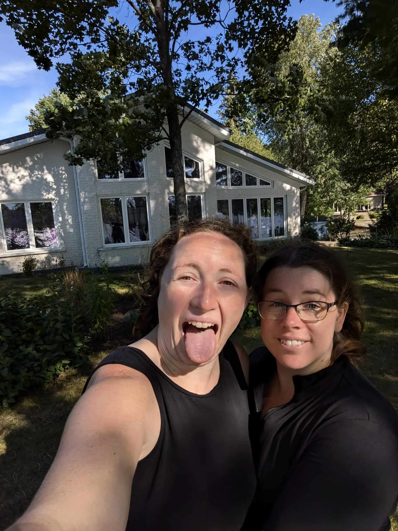 Two women taking a selfie outdoors in front of a modern house with large windows, surrounded by trees and greenery on a sunny day.