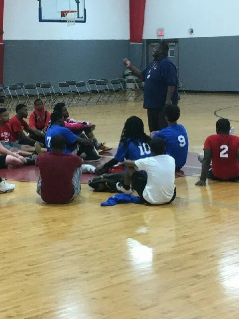 A coach or instructor speaking to a group of young basketball players seated on the gym floor during a practice or meeting.