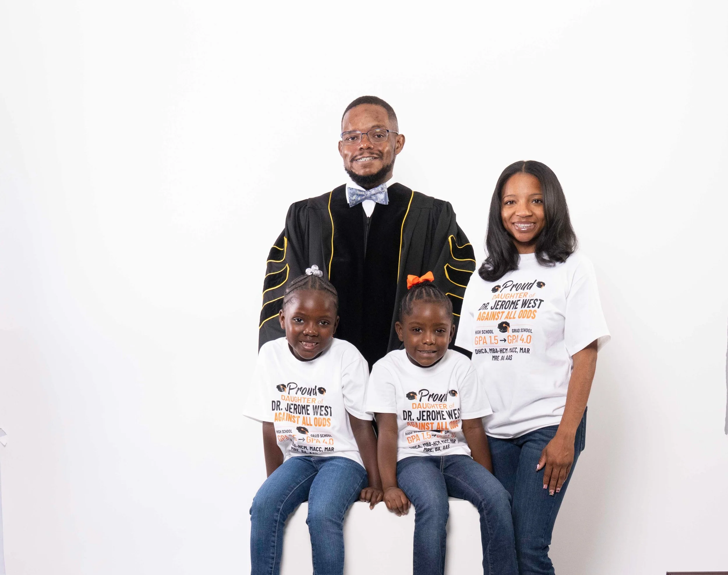 Family portrait of a man in academic regalia, a woman, and two young girls wearing matching T-shirts, standing against a white background.