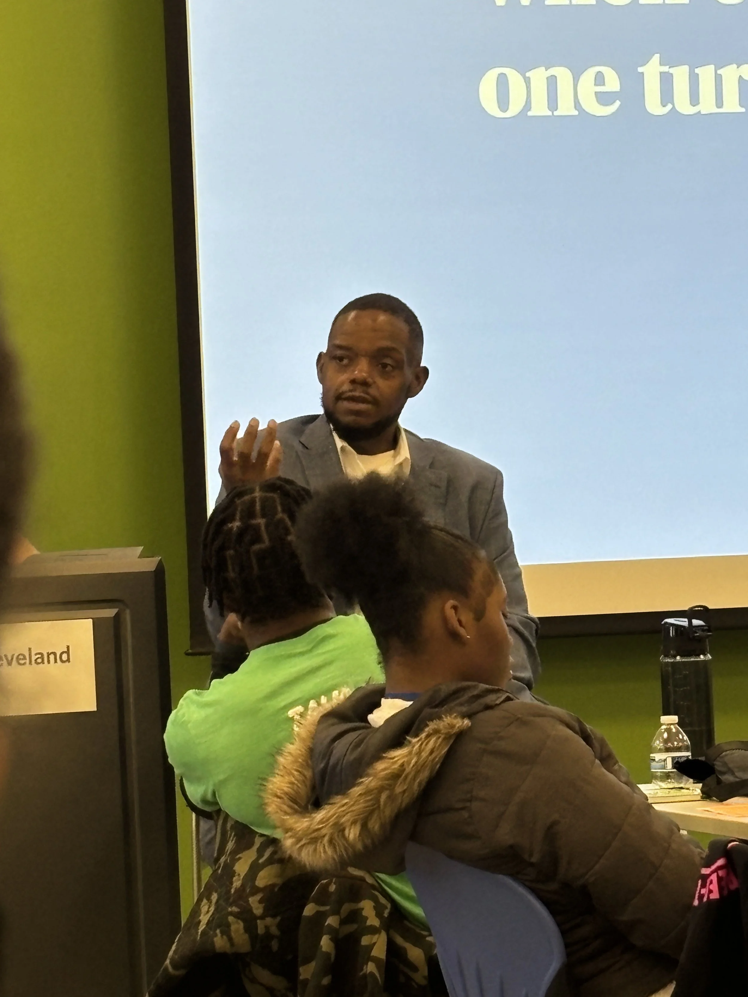 A man in a gray suit and white shirt speaking to an audience in a classroom or conference room, with students sitting at desks listening.