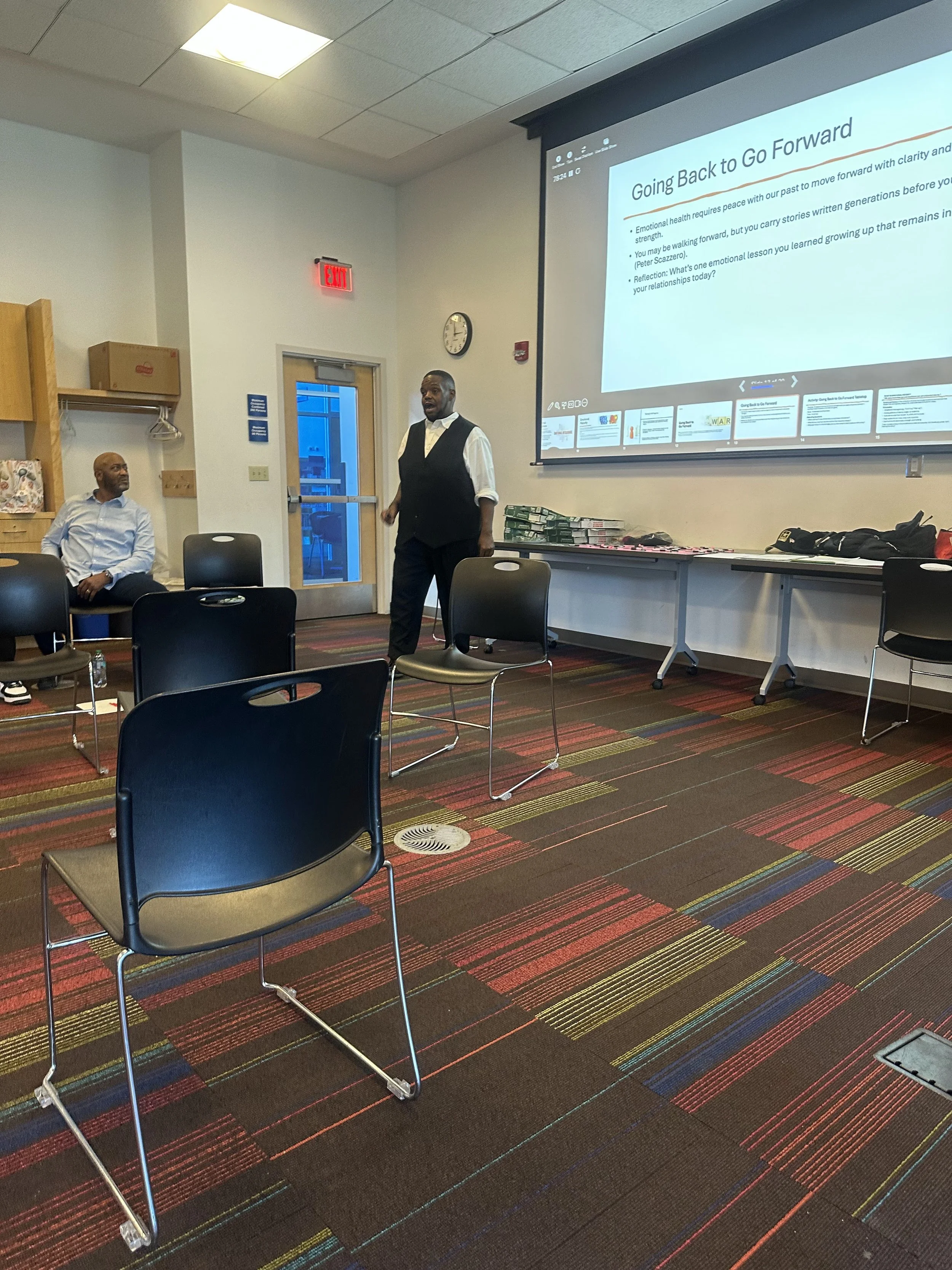 A presentation in progress in a conference room with two men seated and one man standing near a large projection screen displaying a slide titled 'Going Back to Go Forward'. The room has a colorful carpet, chairs, and tables with materials.