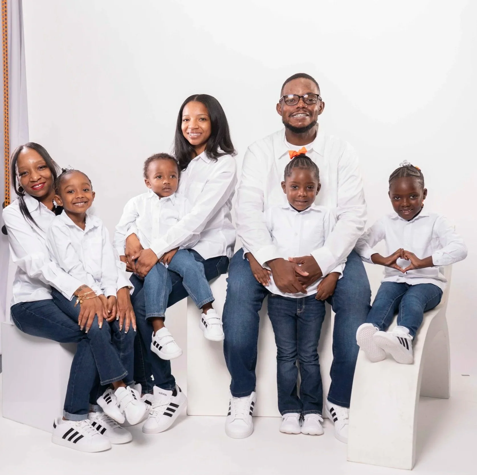 Family portrait of seven members, including two adults and five children, all dressed in white shirts and jeans, sitting and standing on a white abstract bench in a studio with a plain white background.