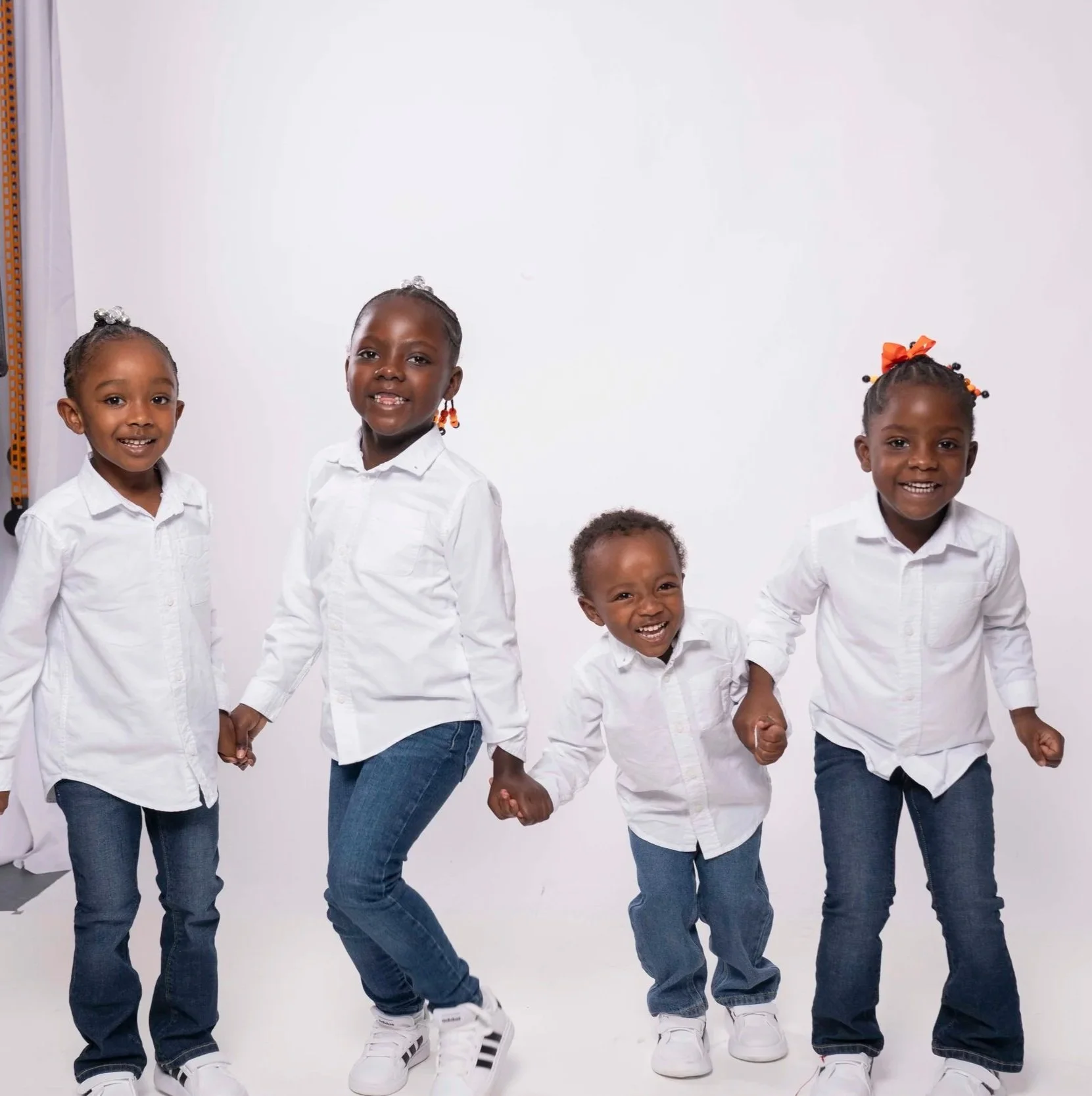Four children holding hands and smiling in a studio with a white background, wearing white shirts and jeans.