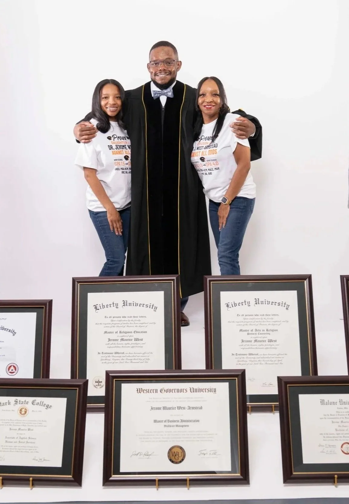 A man in a graduation gown standing between two women, with graduation certificates displayed in front. The women are wearing white t-shirts.