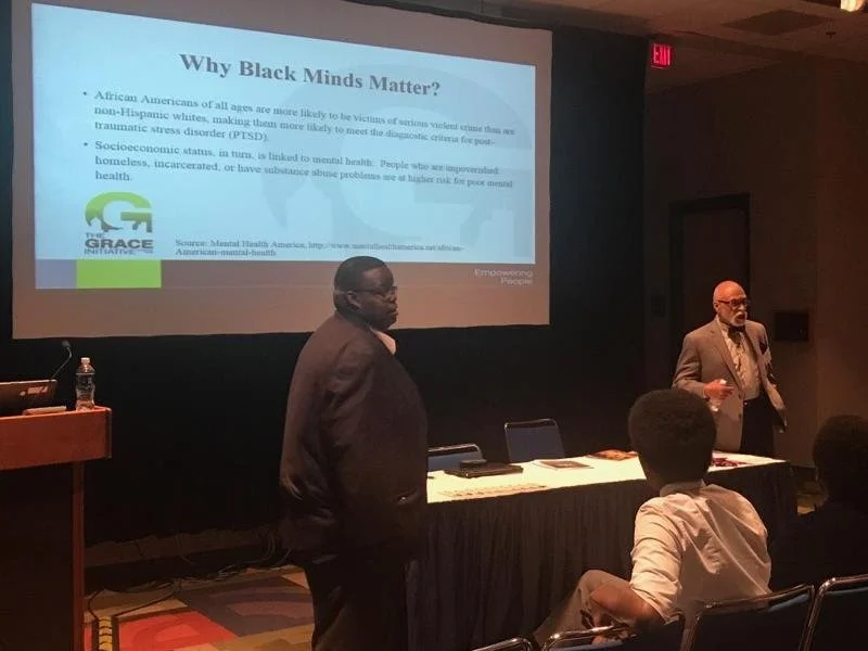 Two men are giving a presentation in a conference room, with a large screen displaying a slideshow titled 'Why Black Minds Matter?' and an audience seated facing them.