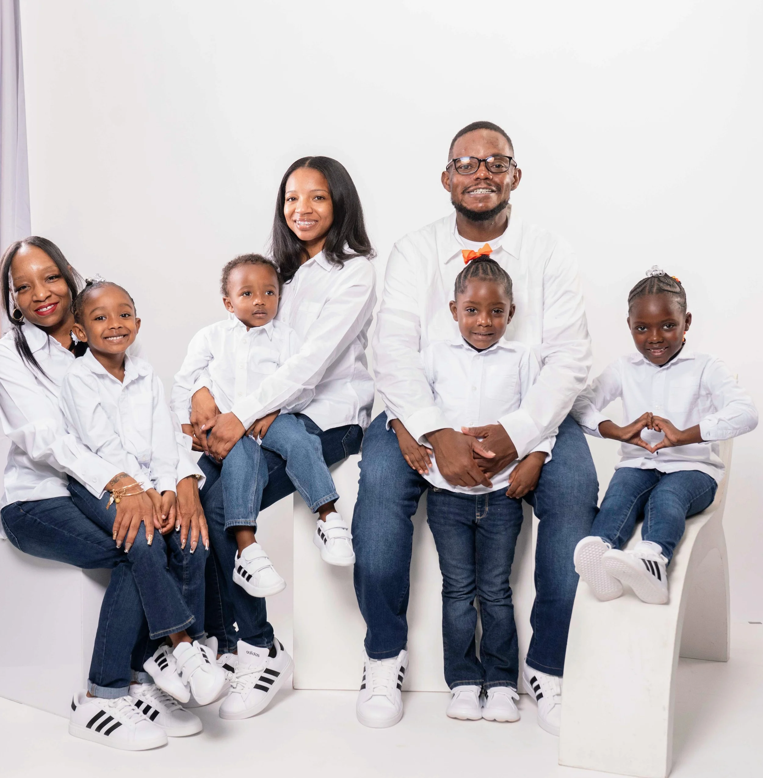 Family portrait of eight people, including two adults and six children, all dressed in white shirts and jeans, sitting and standing against a white background.