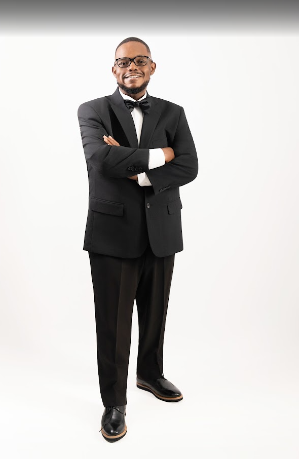 A man in a black tuxedo with a bow tie, glasses, and polished shoes standing against a plain white background, smiling with arms crossed.