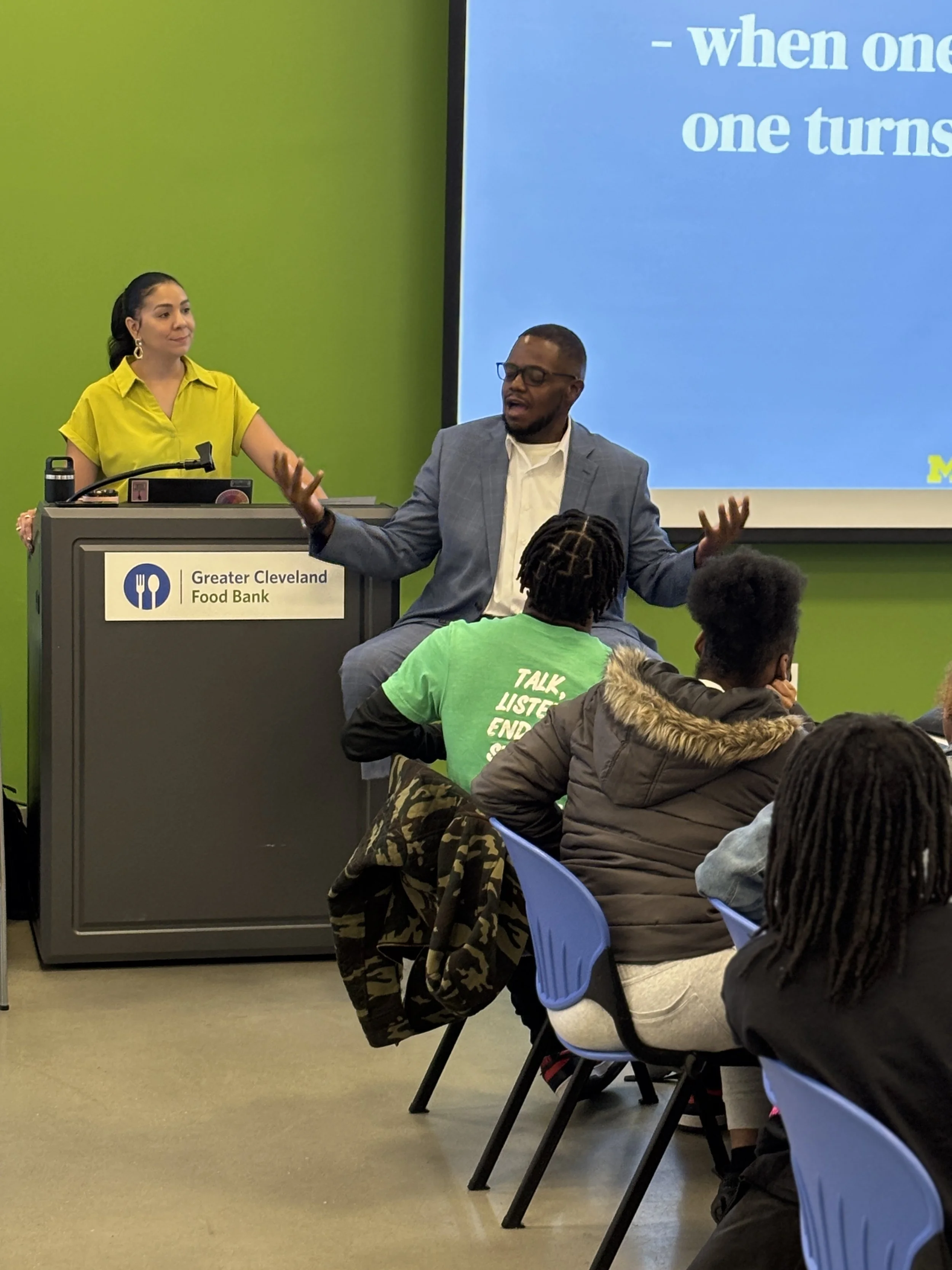 A man giving a presentation in a classroom, with a woman at a podium and an audience of students. The classroom has a green wall and a screen displaying text.