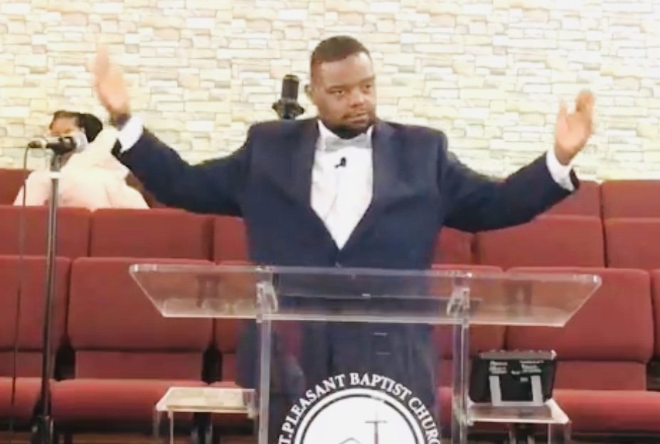 A man in a suit stands behind a clear lectern inside a church, raising his hands during a sermon or speech. The background shows red pews and other attendees.